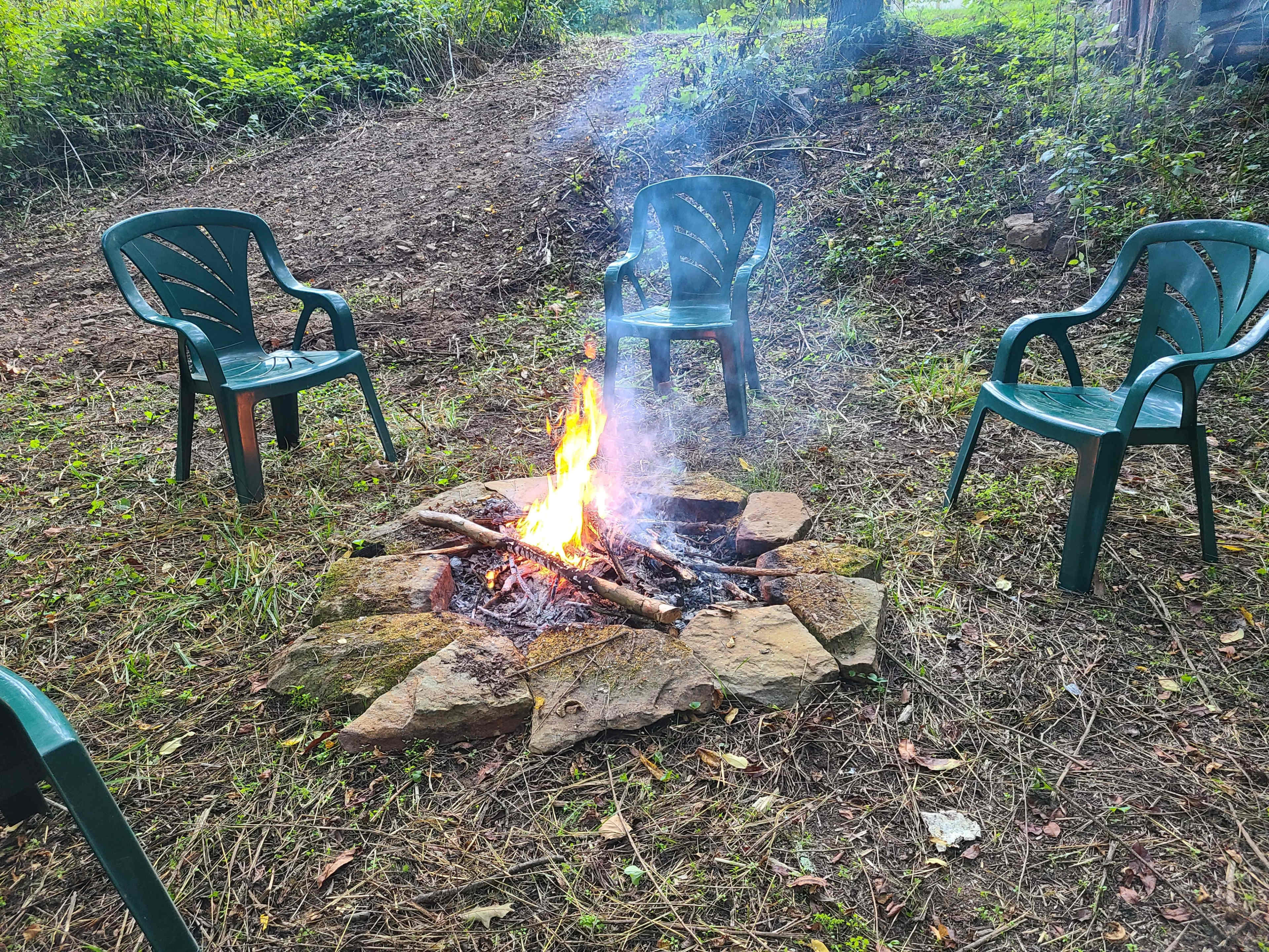 A small campfire burns in a stone circle surrounded by four green chairs in a wooded area.