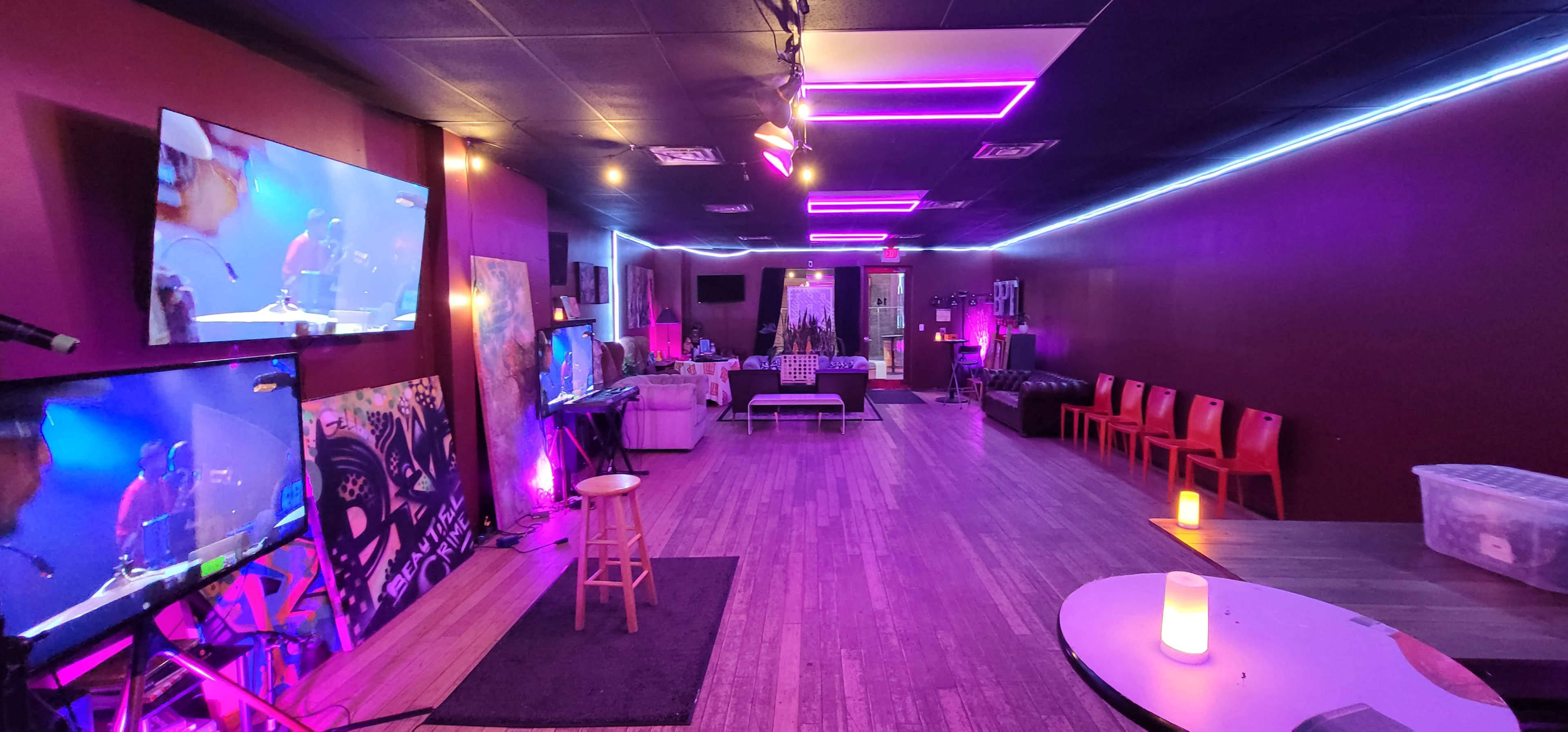The image shows a dimly lit lounge area featuring colorful LED lighting, multiple screens, seating arrangements, and a small stage for performances.
