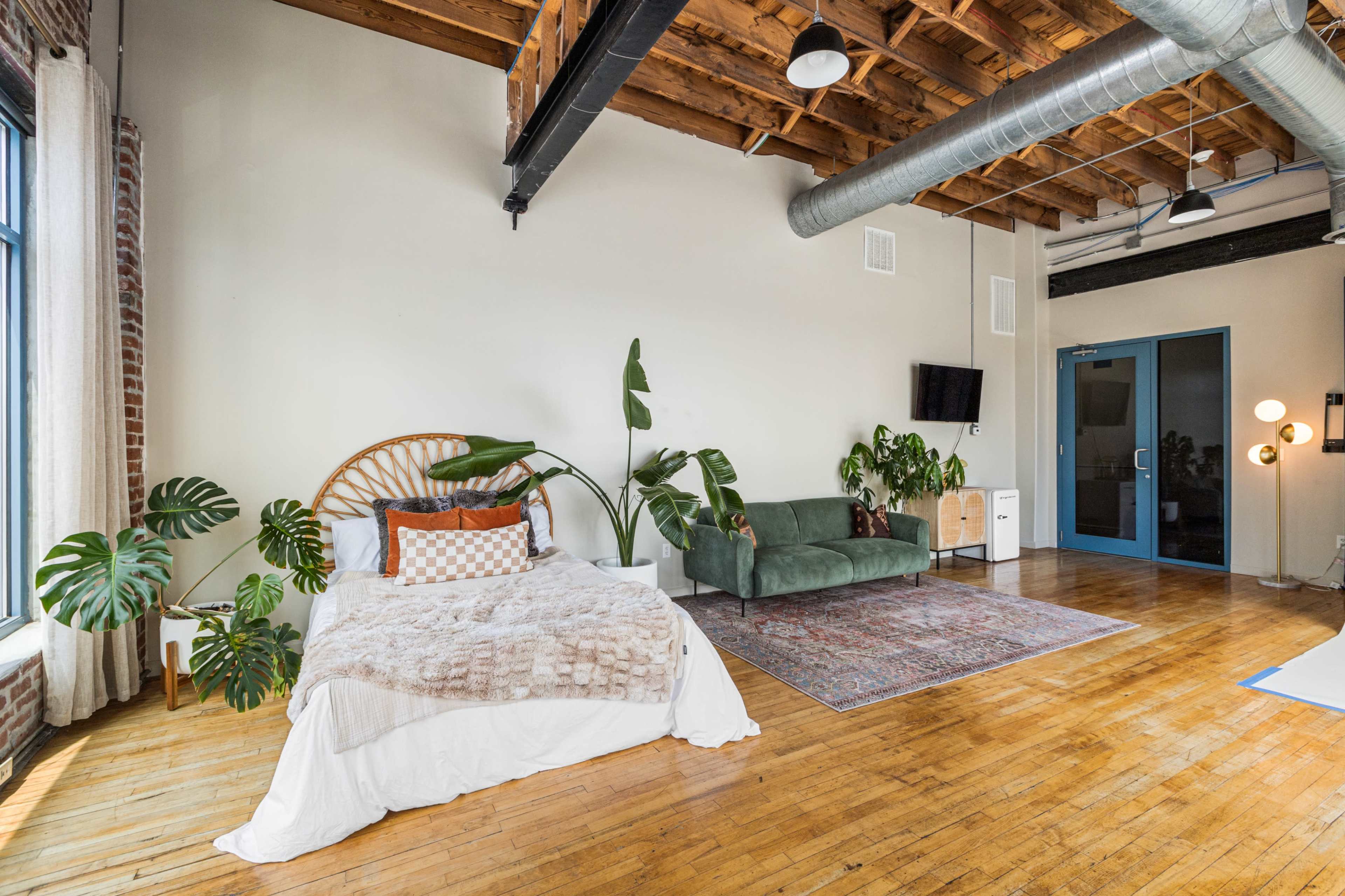 The image shows a modern loft-style room with a bed featuring a rattan headboard, a green sofa, a large area rug, and several potted plants, all under exposed wooden beams and industrial-style lighting.