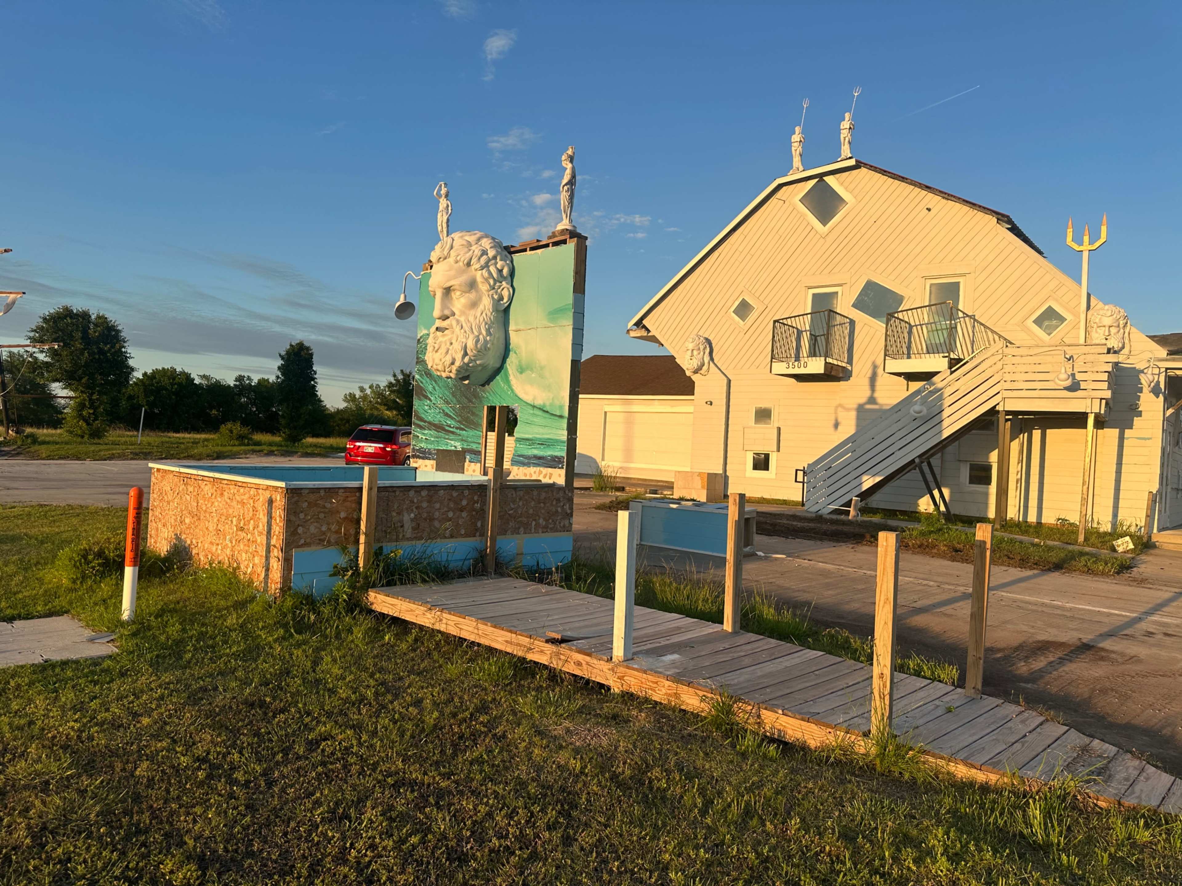 The image shows a distinctive building featuring a mural of a large face, surrounded by sculptures and a wooden walkway leading to a parking area.