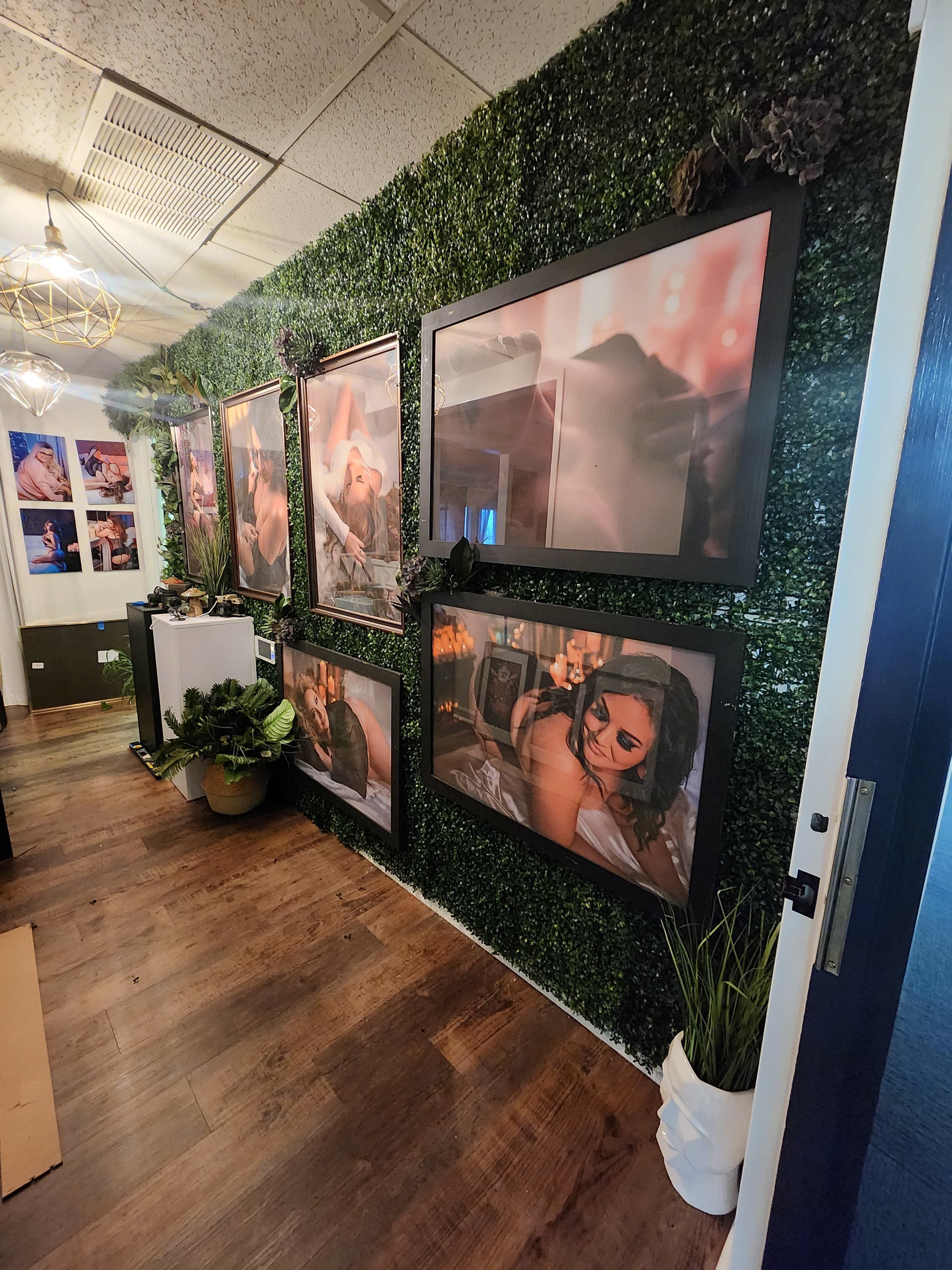 A hallway with a wall covered in artificial green plants, displaying several framed photographs of people.