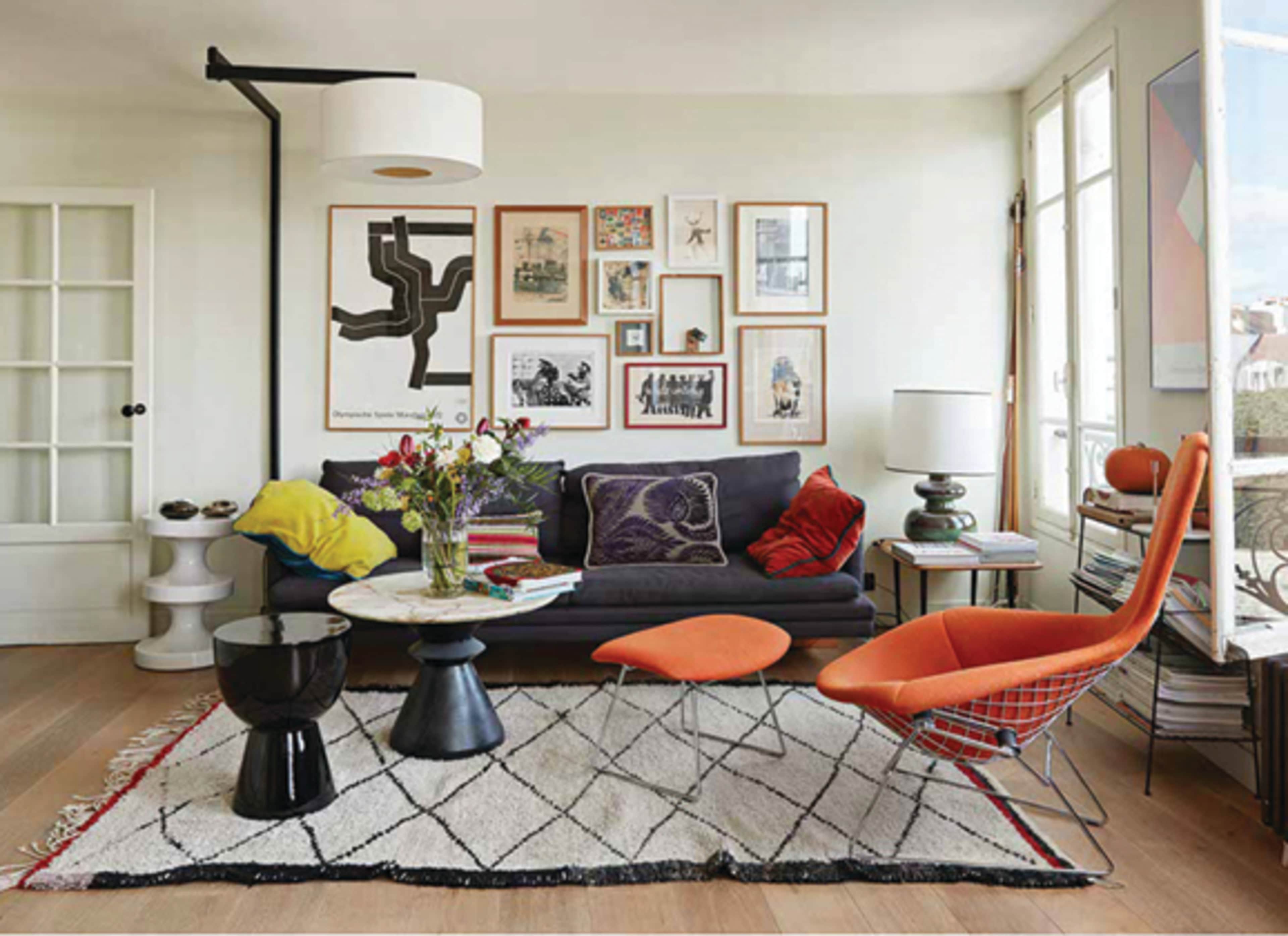 A modern living room features a dark sofa, colorful chairs, a patterned rug, and an assortment of framed artwork on the walls.
