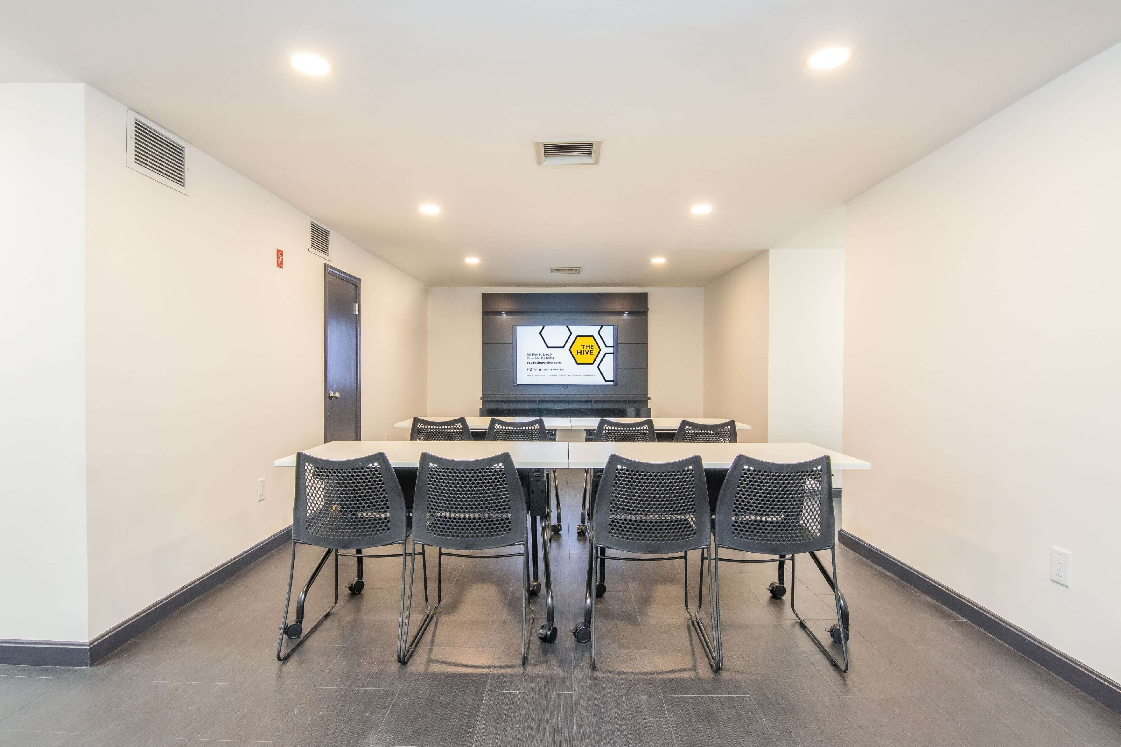 A conference room features a long table surrounded by rolling chairs, with a screen displaying a presentation on one wall.