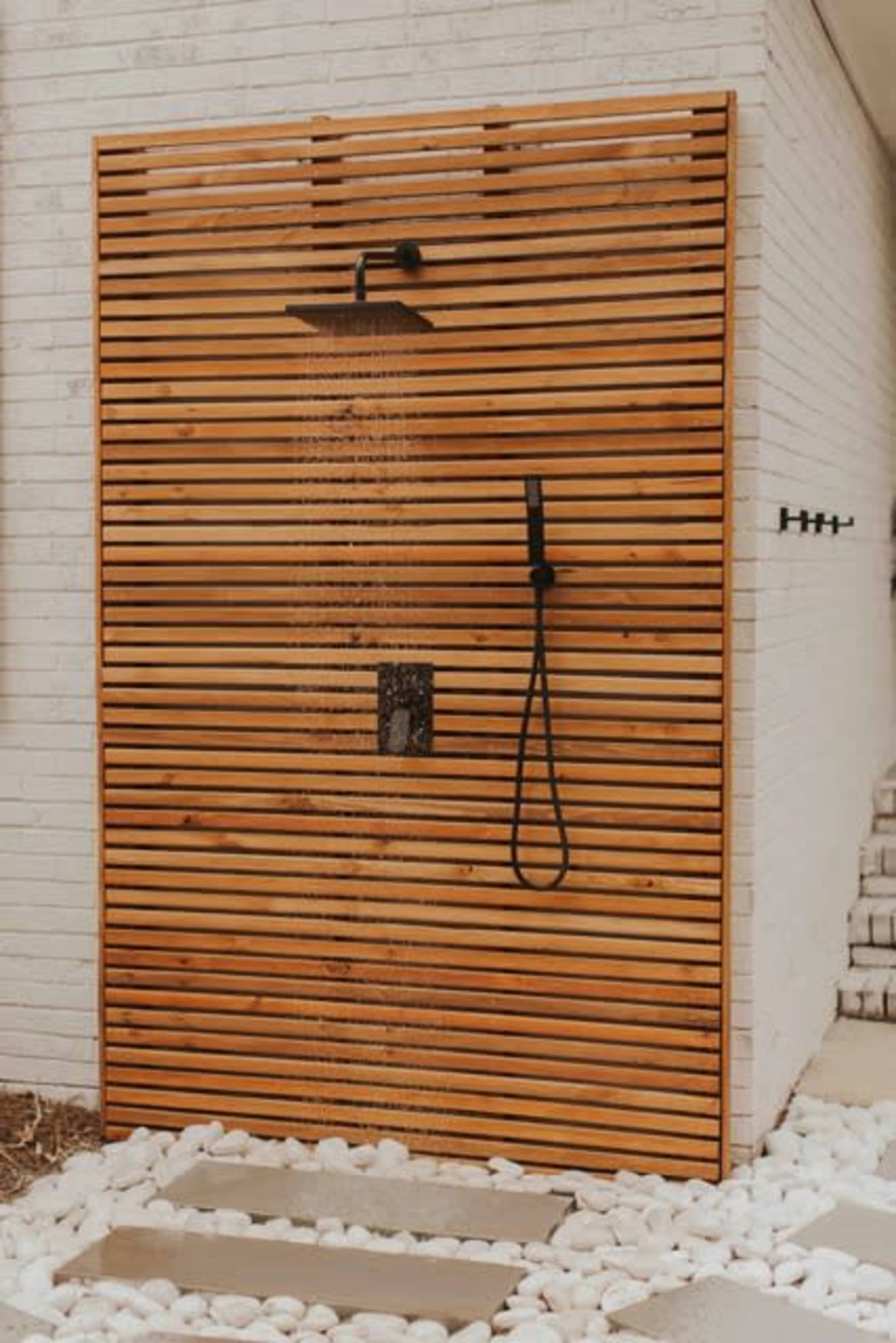 An outdoor shower made of wooden slats with a rain showerhead, situated on a tiled surface surrounded by white pebbles.
