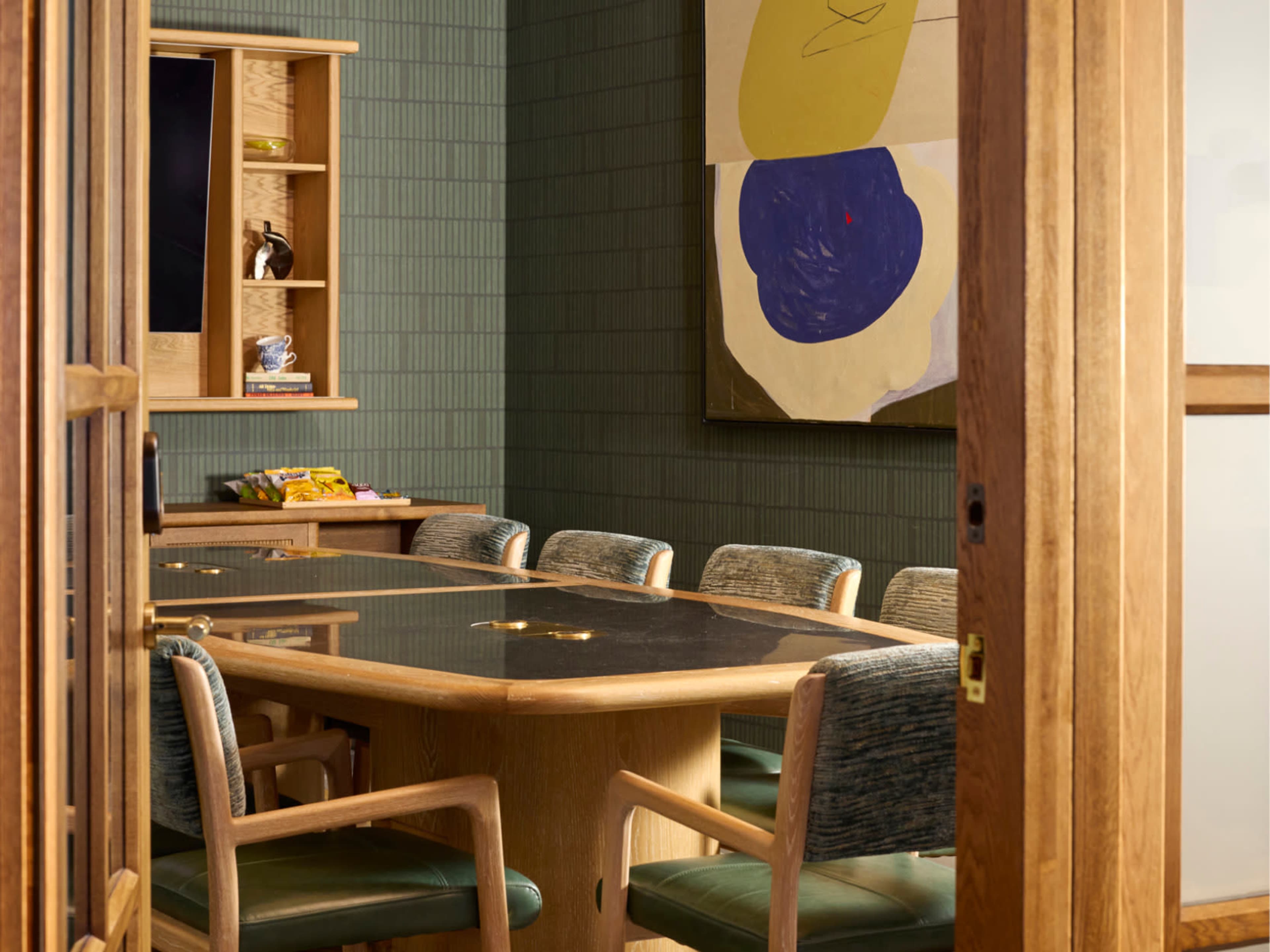 A modern conference room features a round black table surrounded by wooden chairs, with a large abstract painting on the wall and a shelving unit in the background.