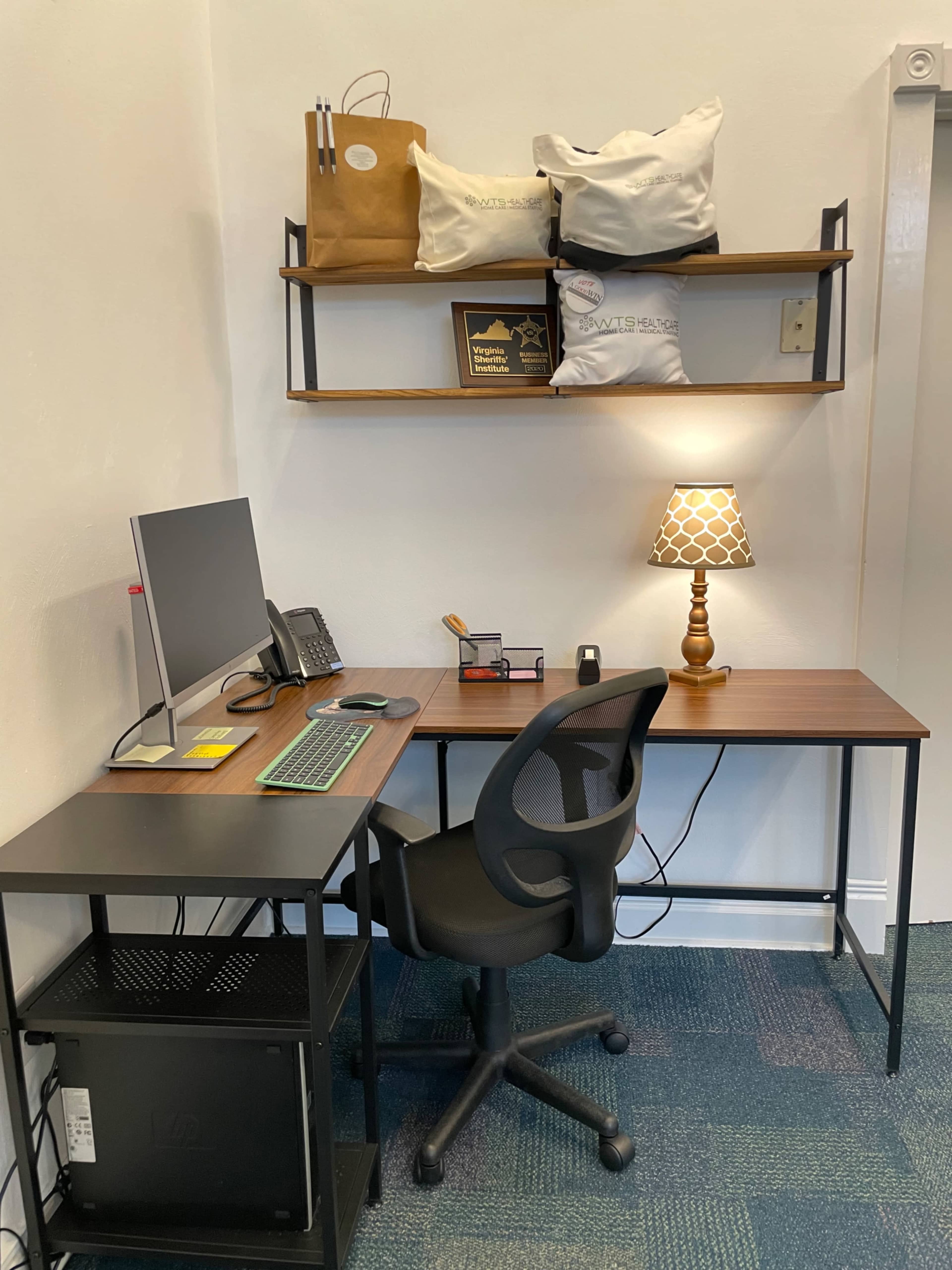 A workspace featuring a desk with a computer, a chair, a lamp, and shelves above with bags and decorative items.