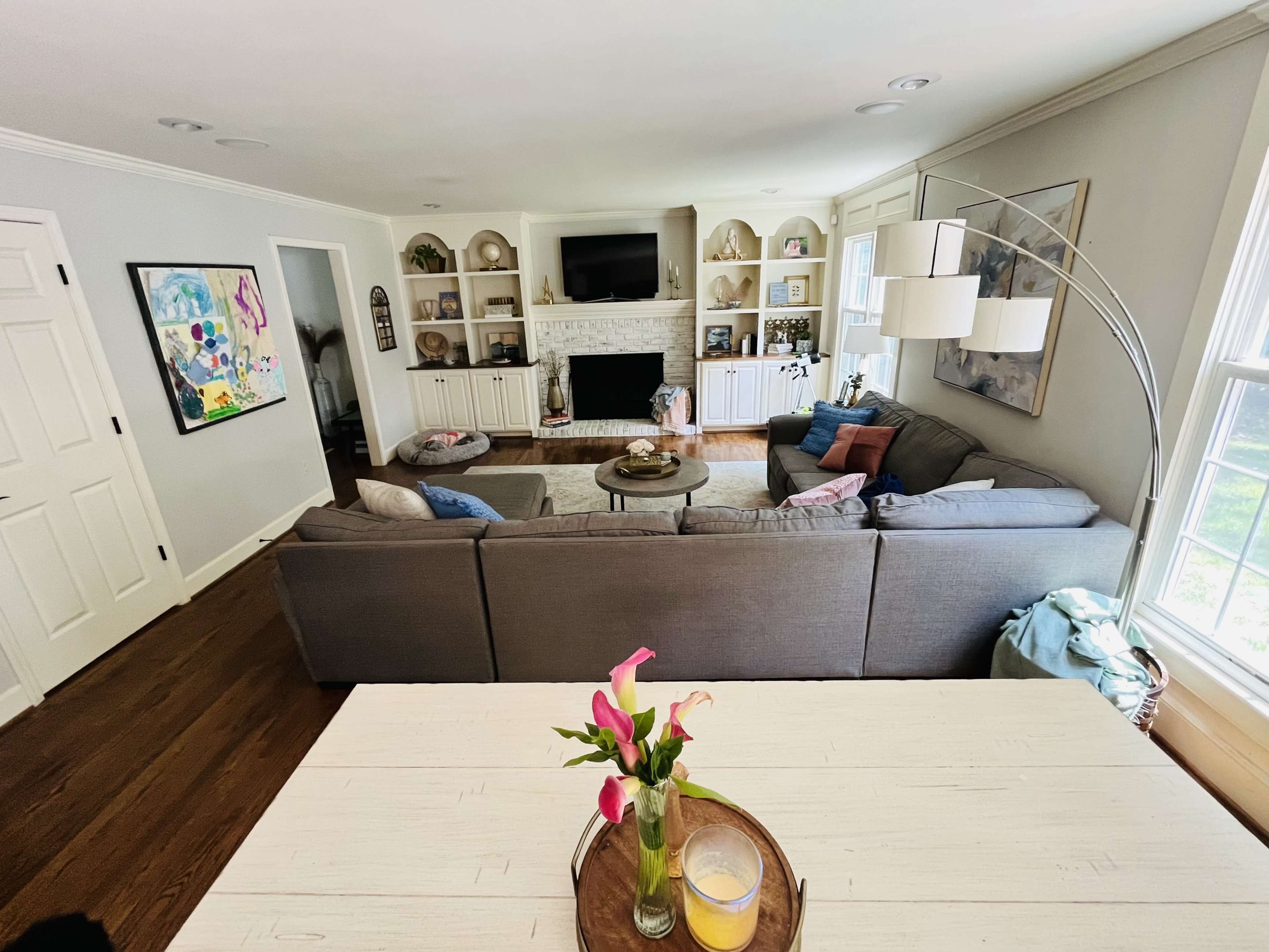 The image shows a spacious living room furnished with a gray sectional sofa, a coffee table, and a dining table with a vase of flowers.