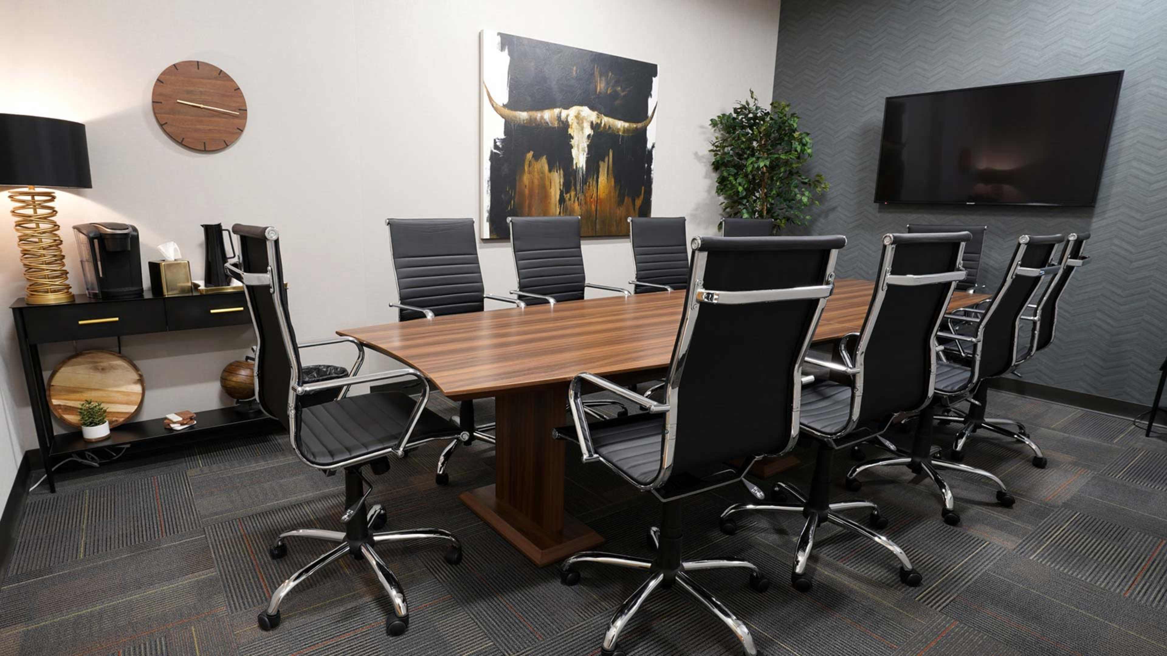 A modern conference room features a long wooden table surrounded by black ergonomic chairs, with a large television and decorative artwork on the walls.