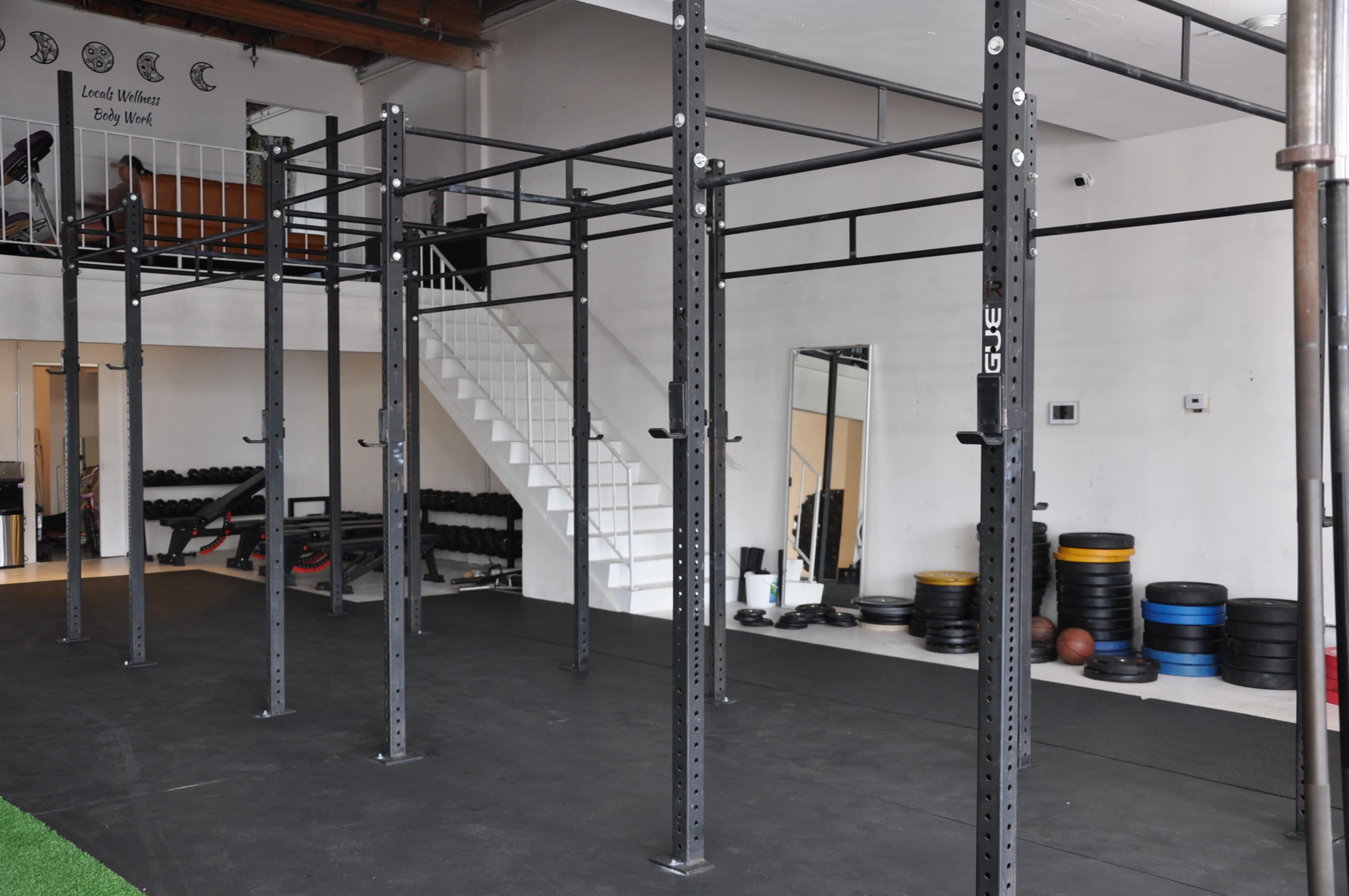 The image shows a gym interior featuring a series of black workout racks and equipment, along with a staircase in the background.