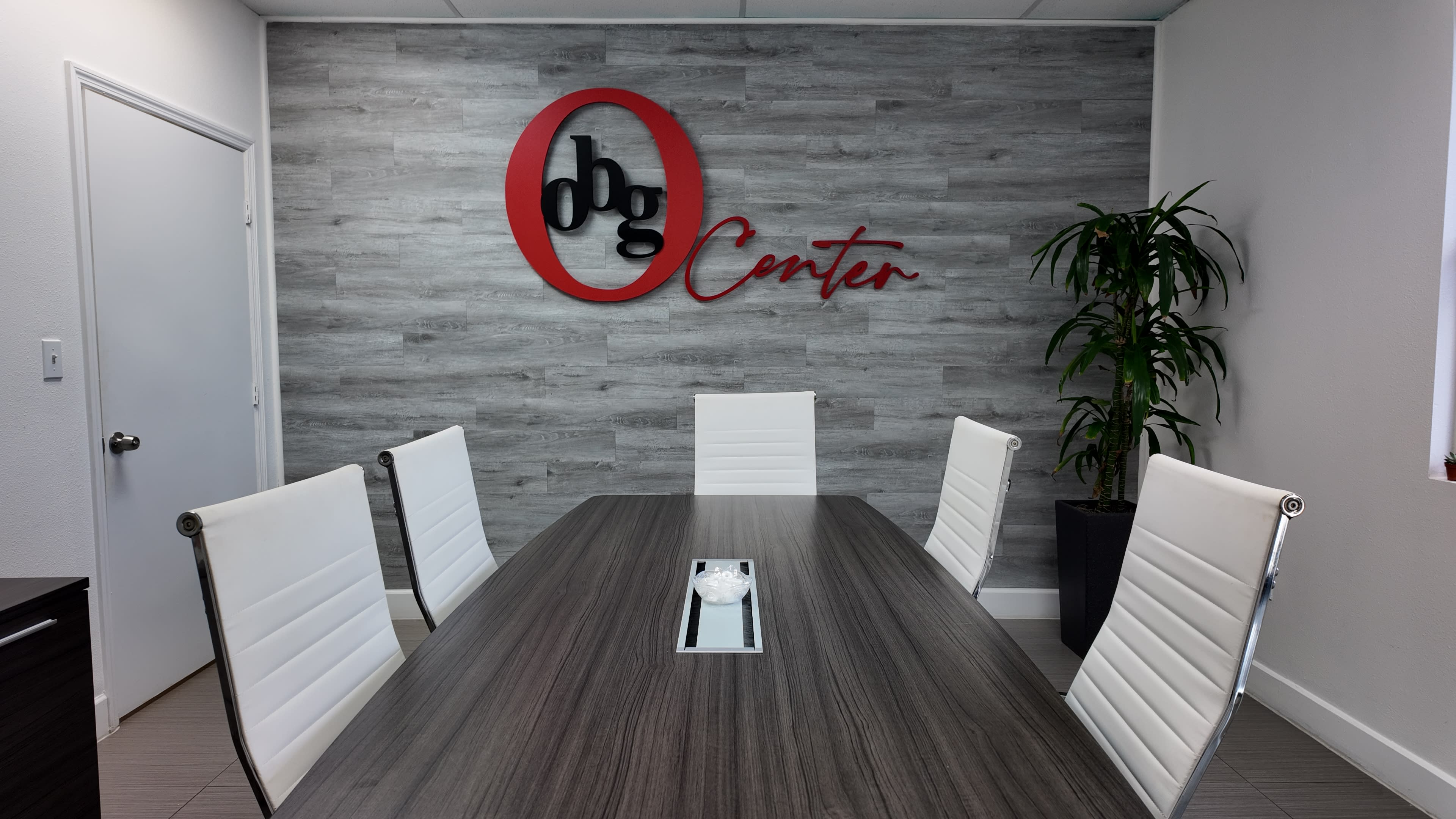 A conference room features a long table surrounded by white chairs, with a large red and black logo on the wall.