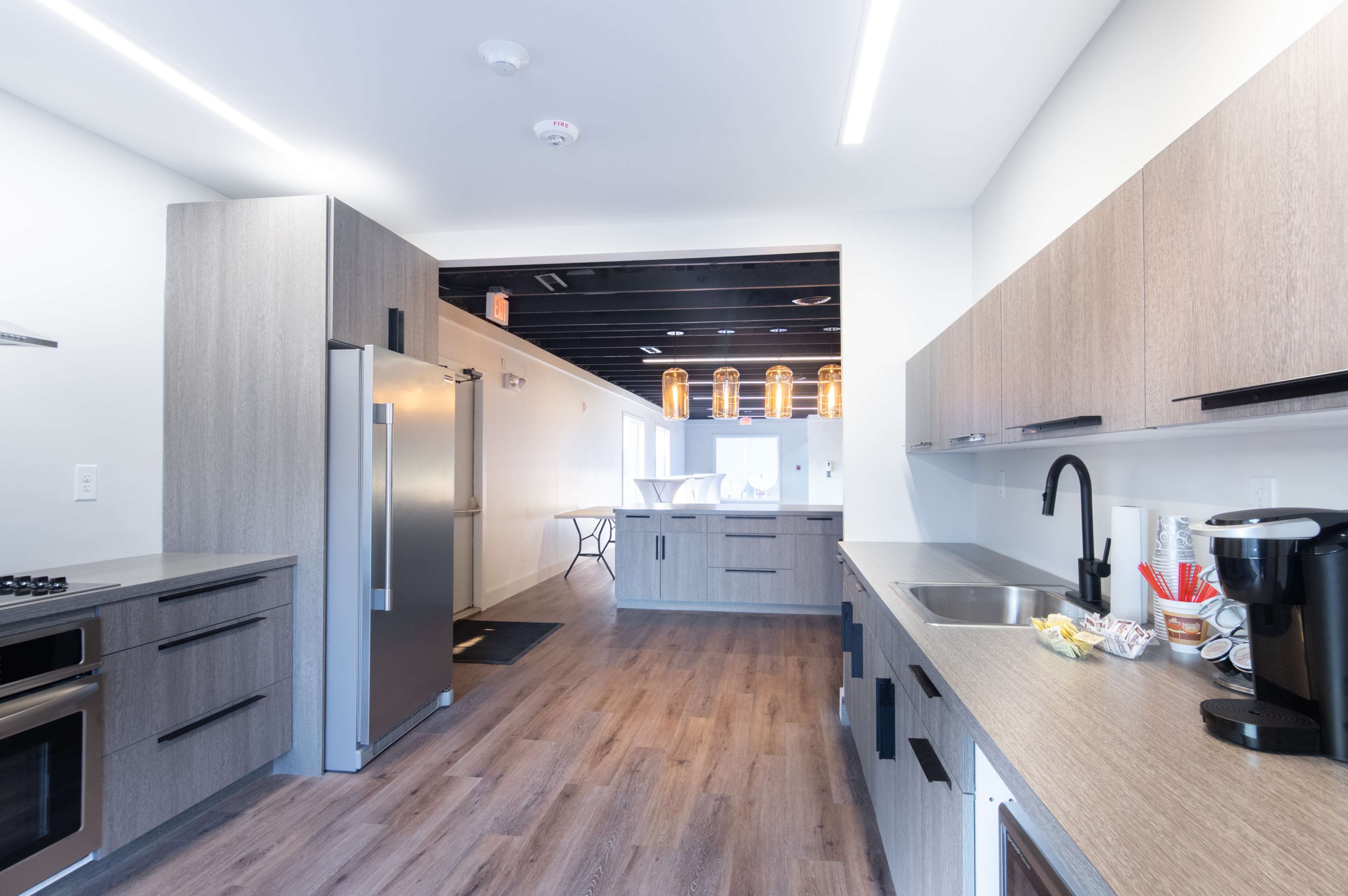 A modern kitchen with grey cabinetry, stainless steel appliances, and a light wood floor, featuring an open layout leading to another room.