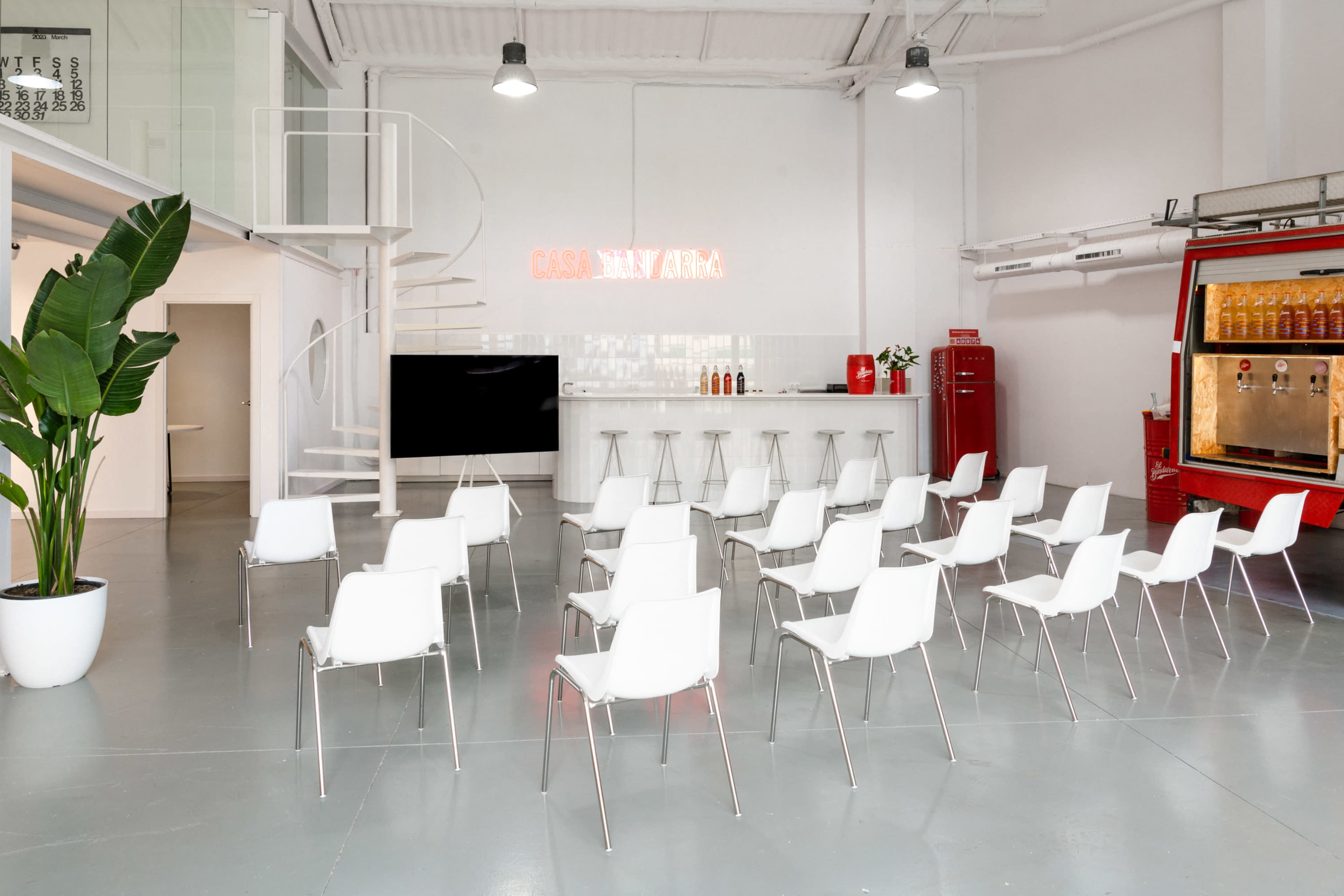 The image shows a modern, minimalistic interior space with white chairs arranged in rows facing a bar area, accompanied by a vibrant red vending machine and a large TV screen.