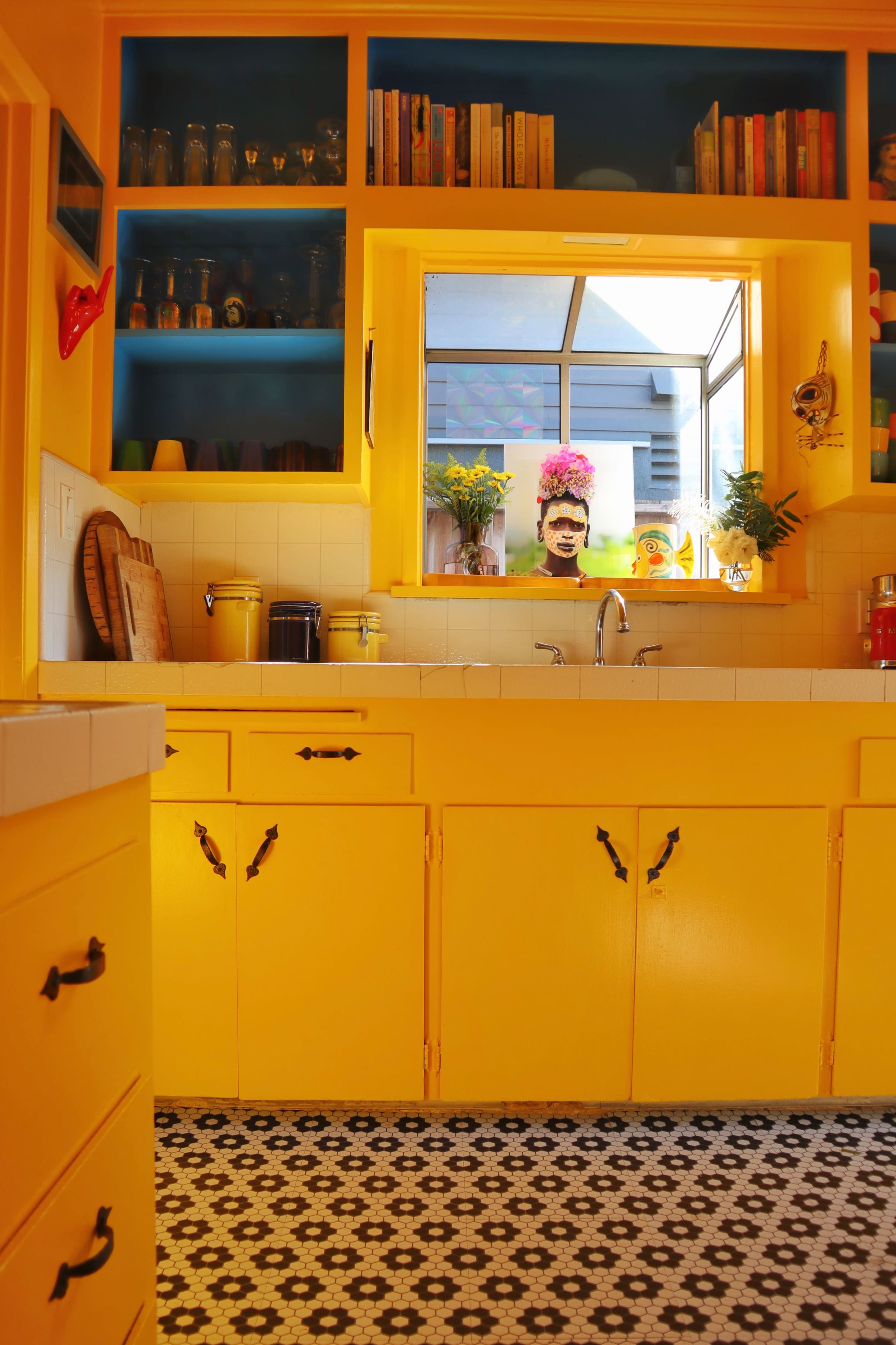 A bright yellow kitchen with open shelves filled with books and glassware, and a window that offers a view of the outdoors.