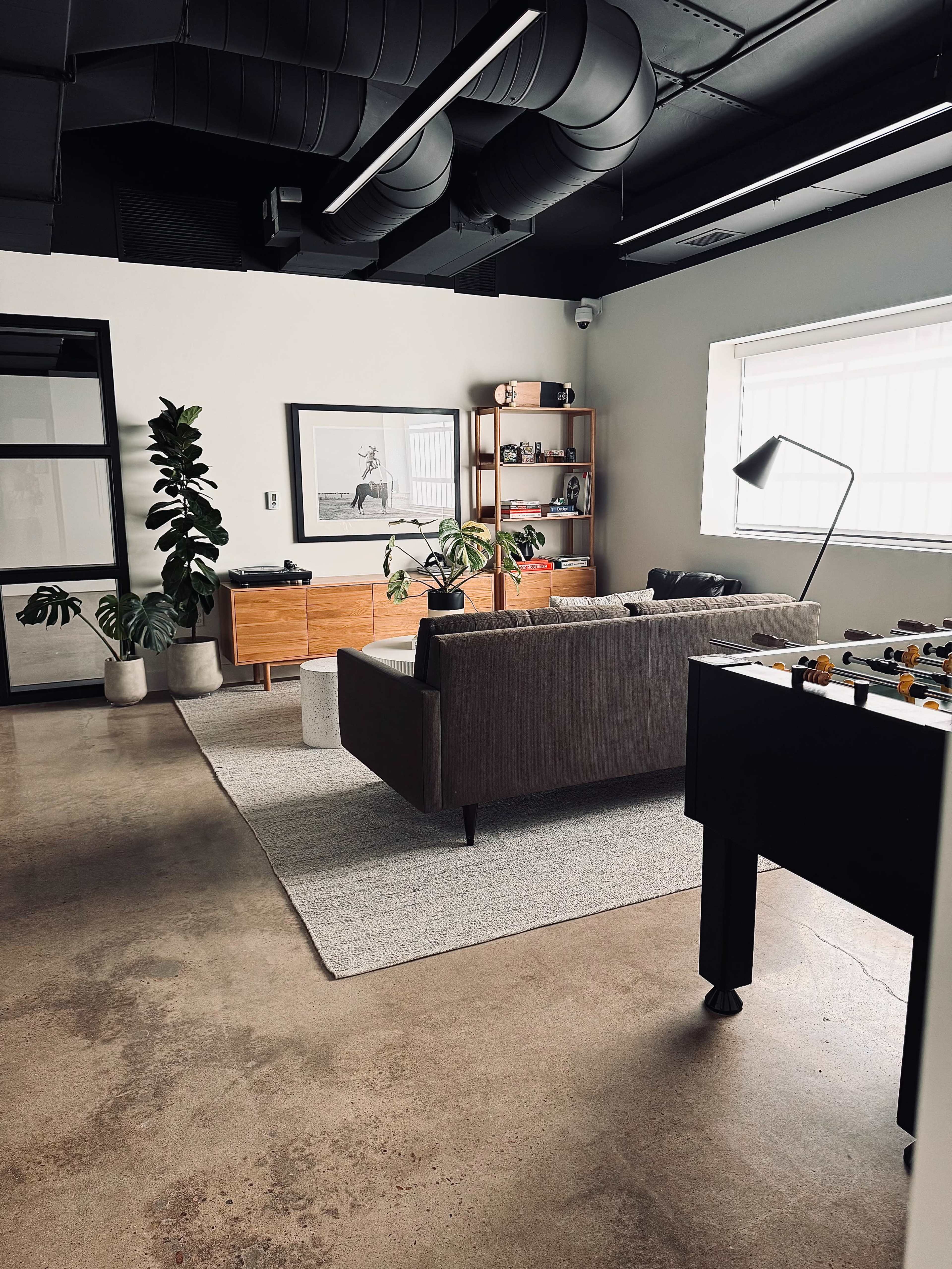 A modern office lounge features a gray sofa, wooden storage unit, large plants, and a foosball table on a concrete floor.