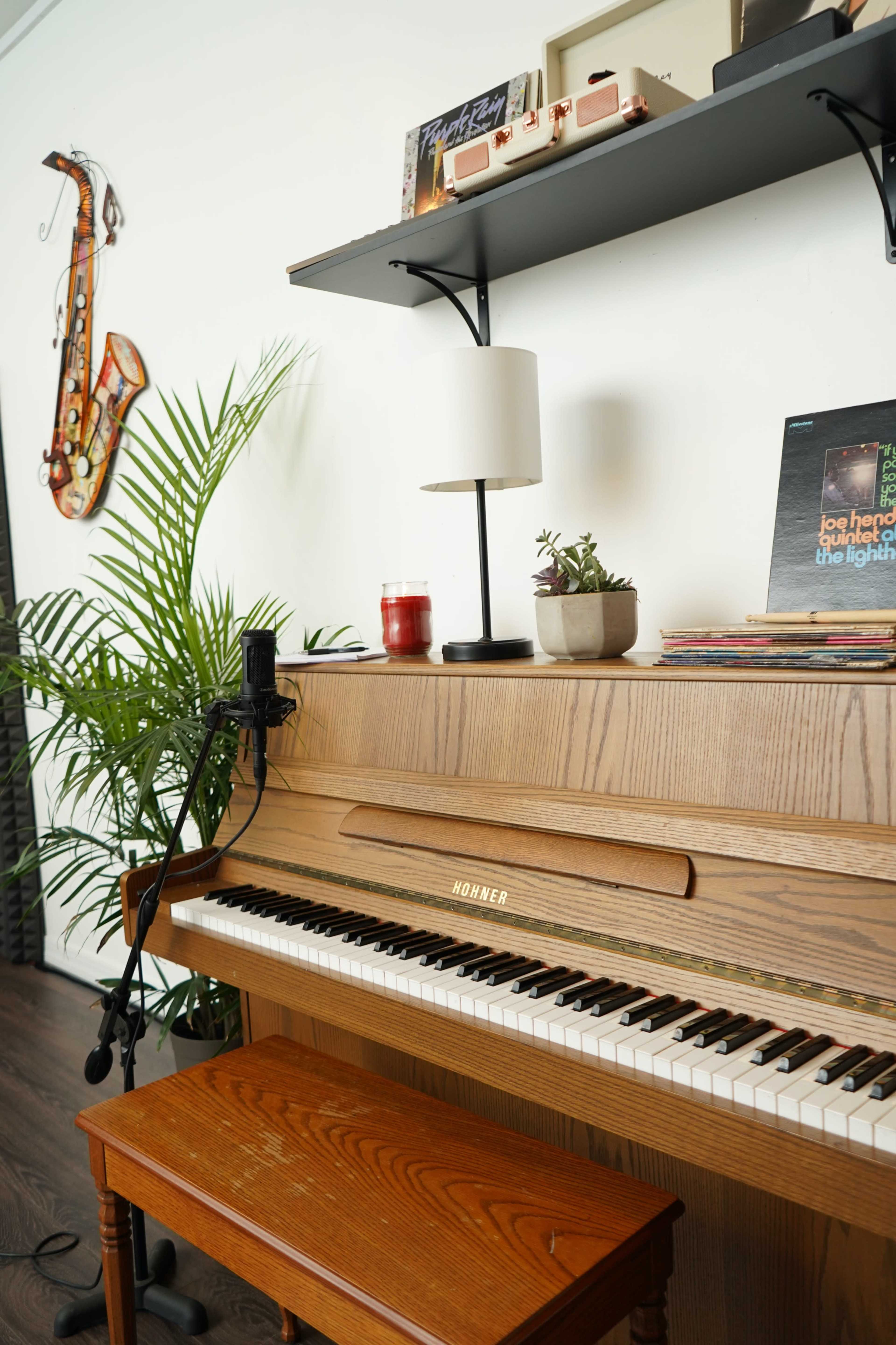 A wooden piano is positioned against a wall, accompanied by a microphone, a lamp, a small plant, and various records on a shelf above.