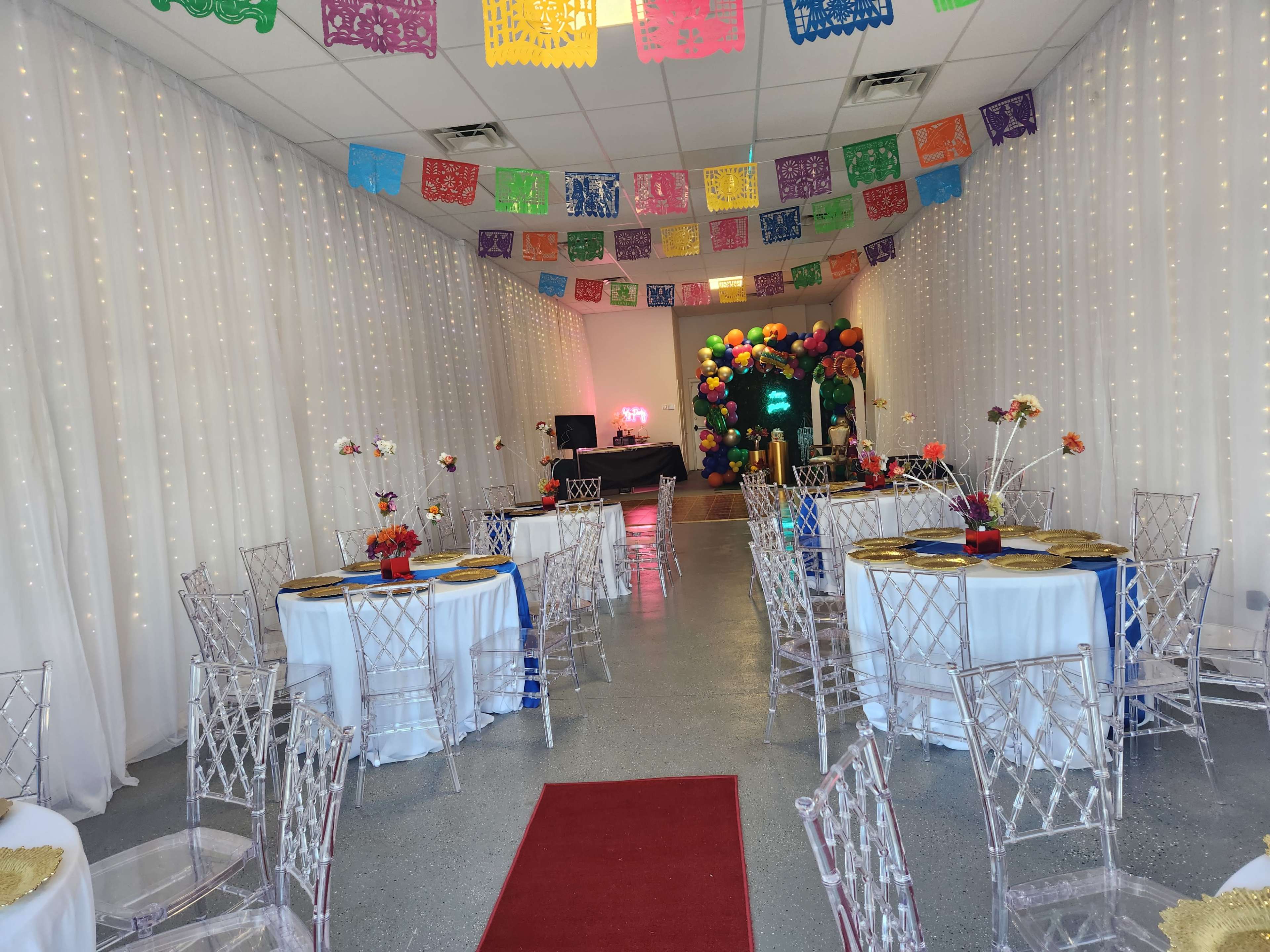A decorated event space with tables covered in white tablecloths, surrounded by transparent chairs, and colorful papel picado banners hanging from the ceiling.