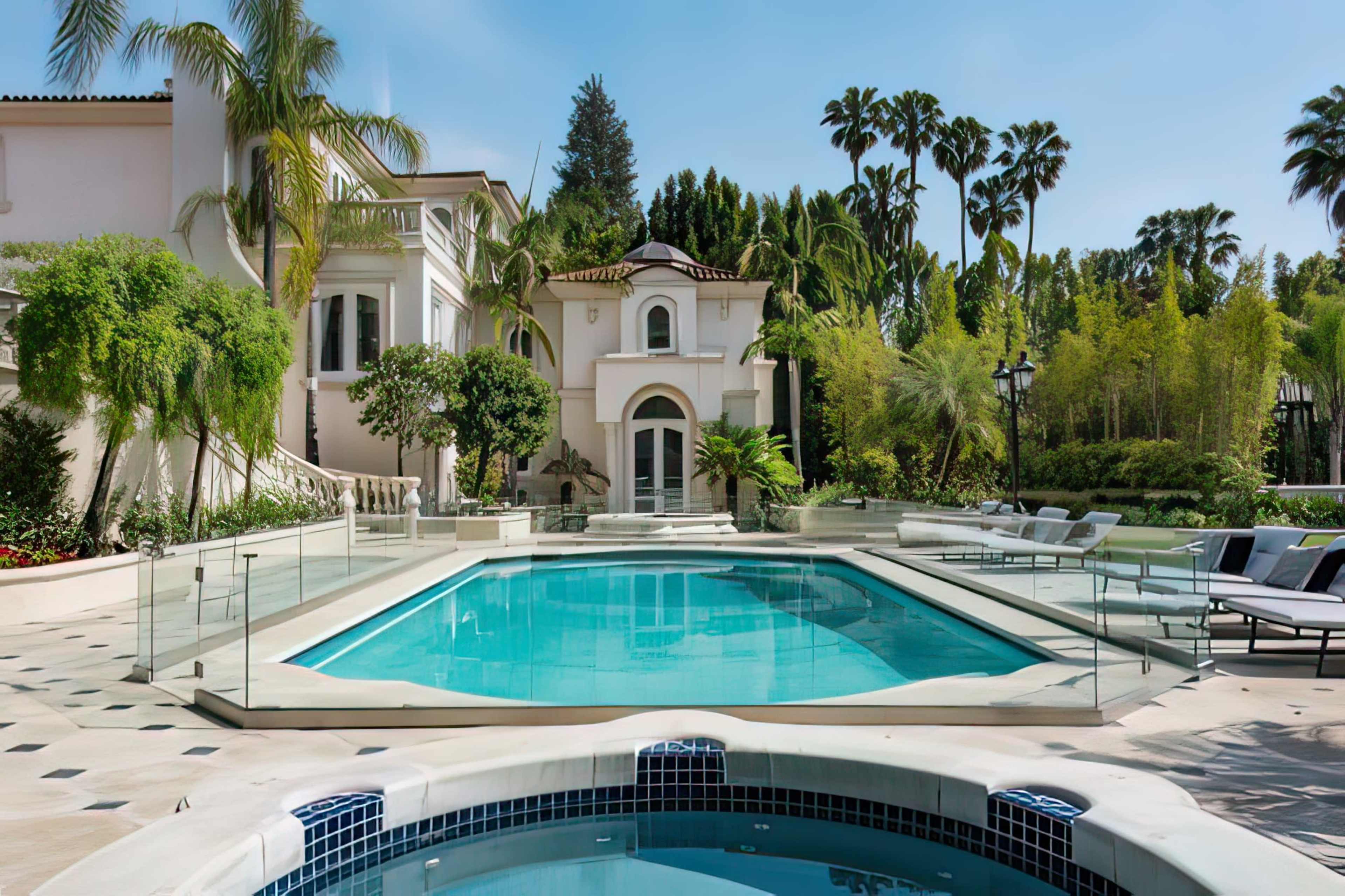A luxury pool area features a large swimming pool surrounded by palm trees and a modern, elegant home.
