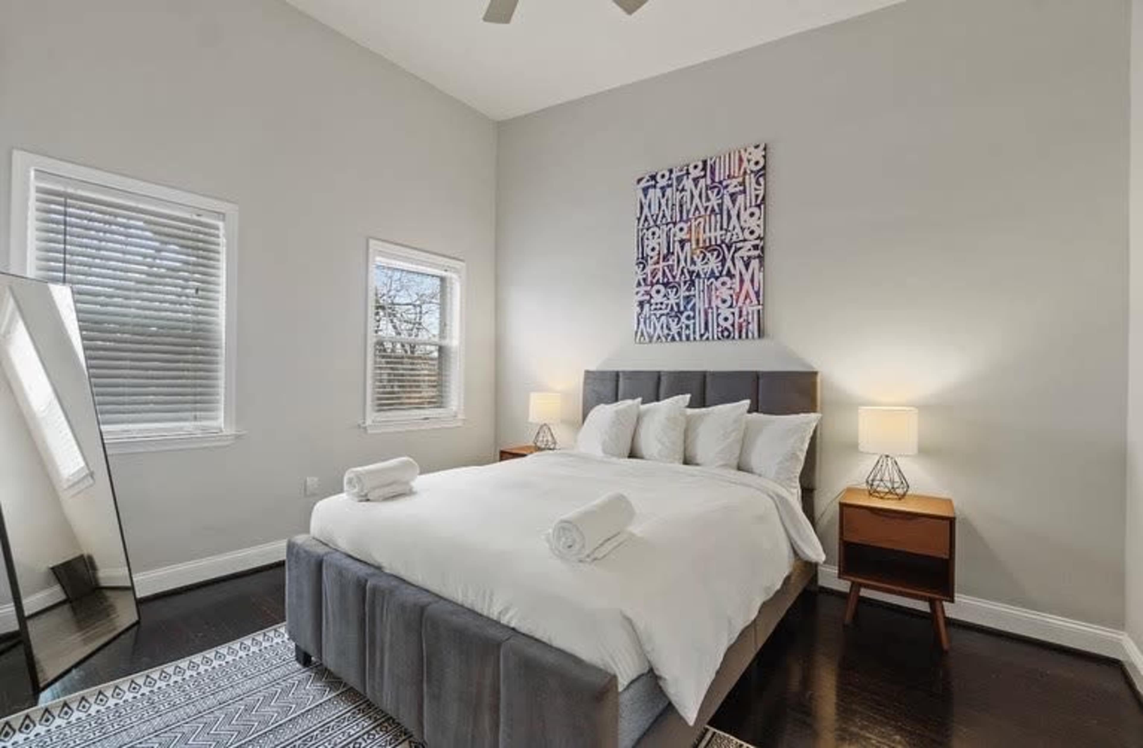 The image shows a neatly organized bedroom featuring a large bed with white bedding, two bedside lamps, and a mirror beside the bed.