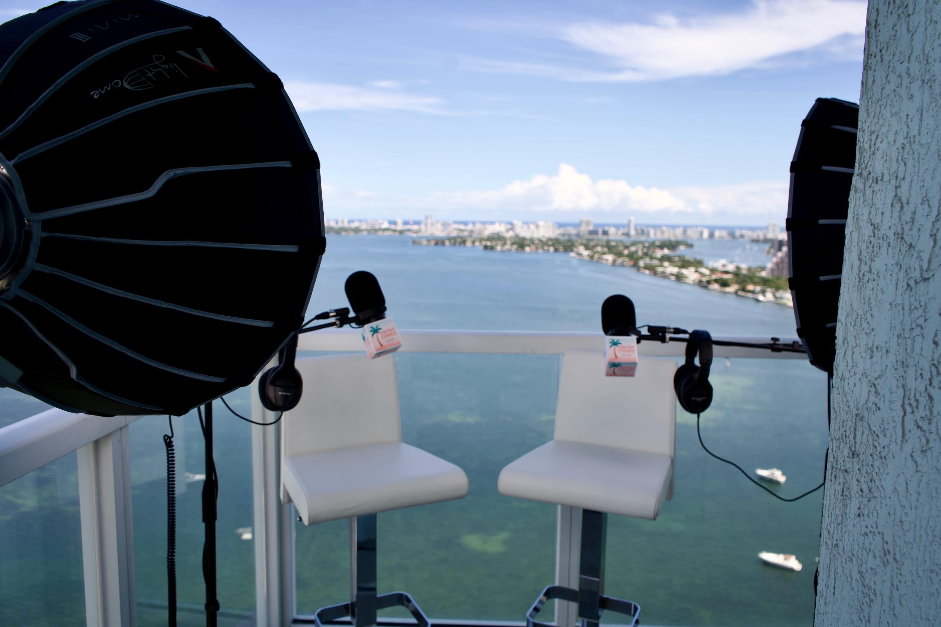 The scene shows two white barstools and microphones set up on a balcony overlooking a body of water and a city skyline in the distance.