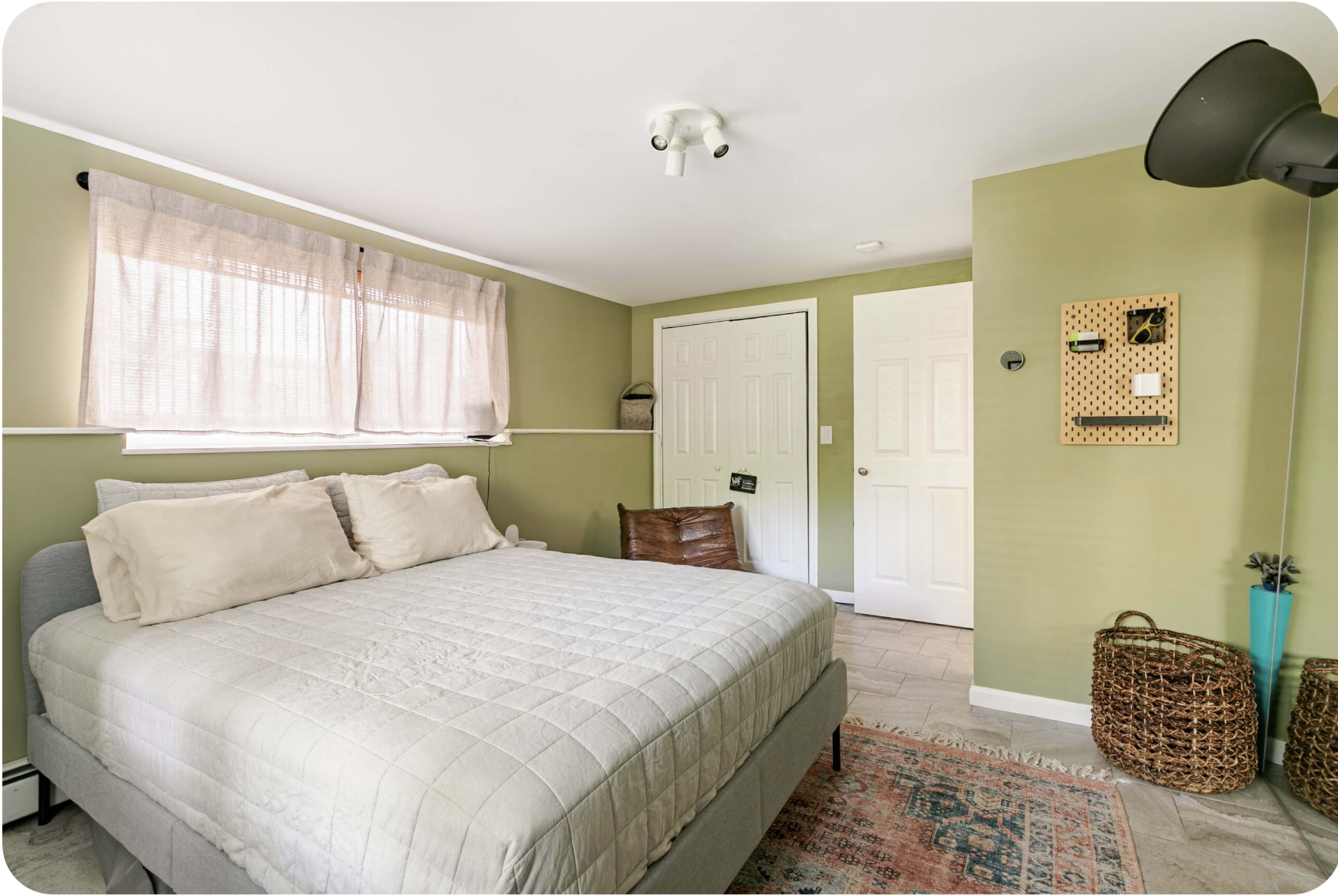 The room features a bed with neutral bedding, a window with sheer curtains, a door, and a woven basket on the floor.