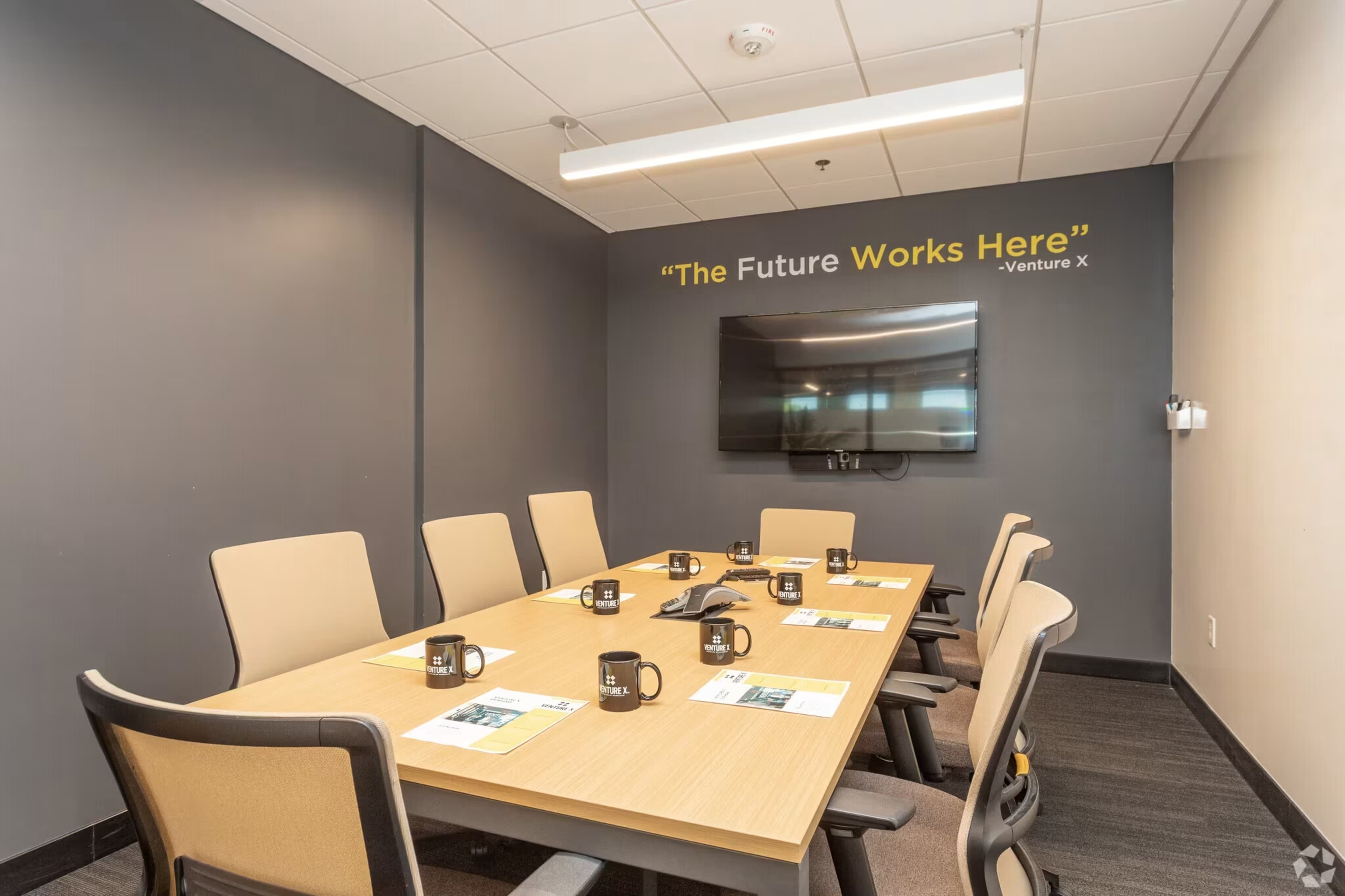 A conference room features a long table with six chairs, a large wall-mounted television, and a motivational quote above.