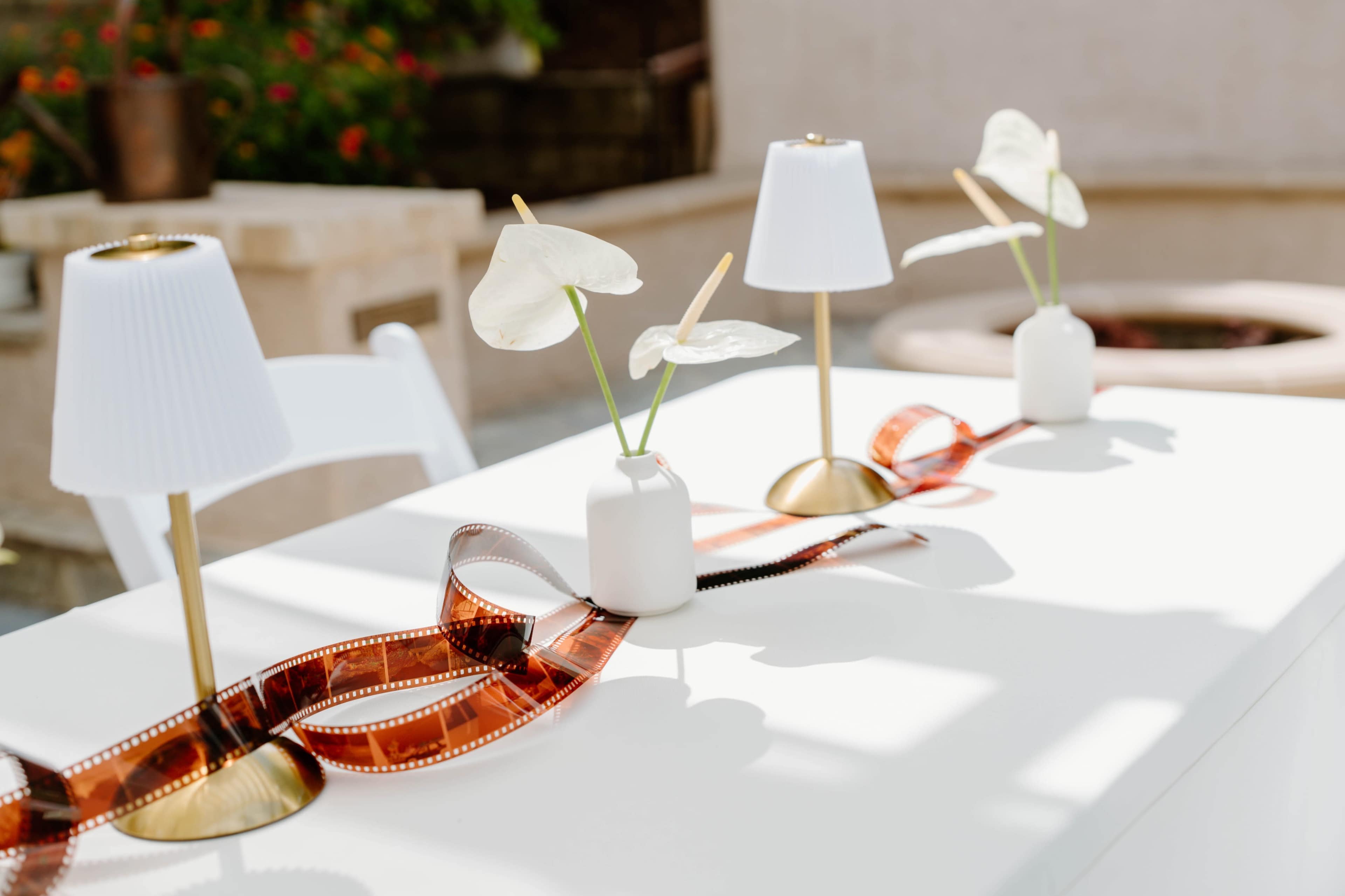 The image shows a tablescape with three decorative table lamps, white vases containing white flowers, and a film strip running along the center.