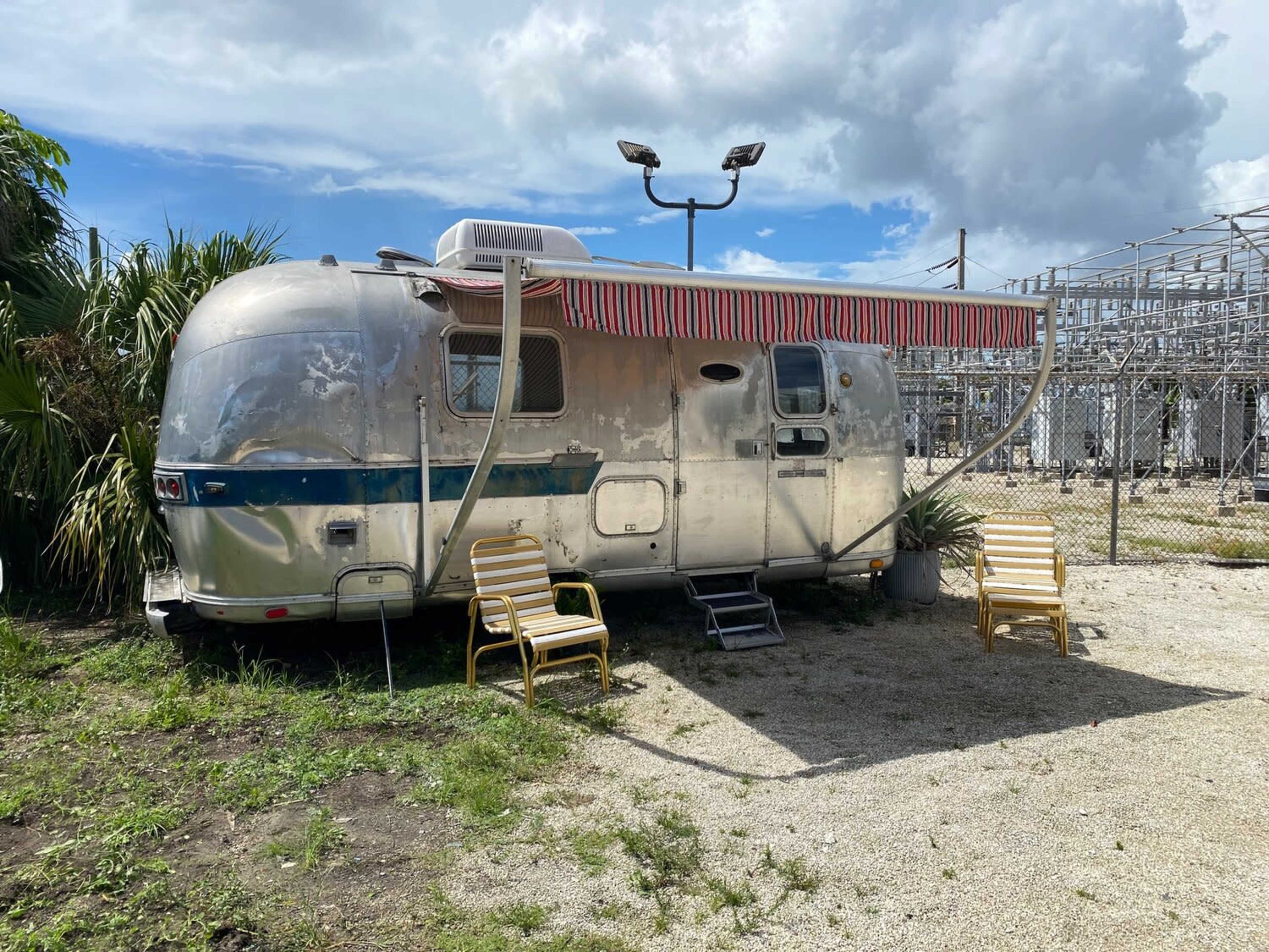 An aluminum Airstream trailer is parked next to a fence, with two yellow chairs positioned outside and overhead power lines in the background.