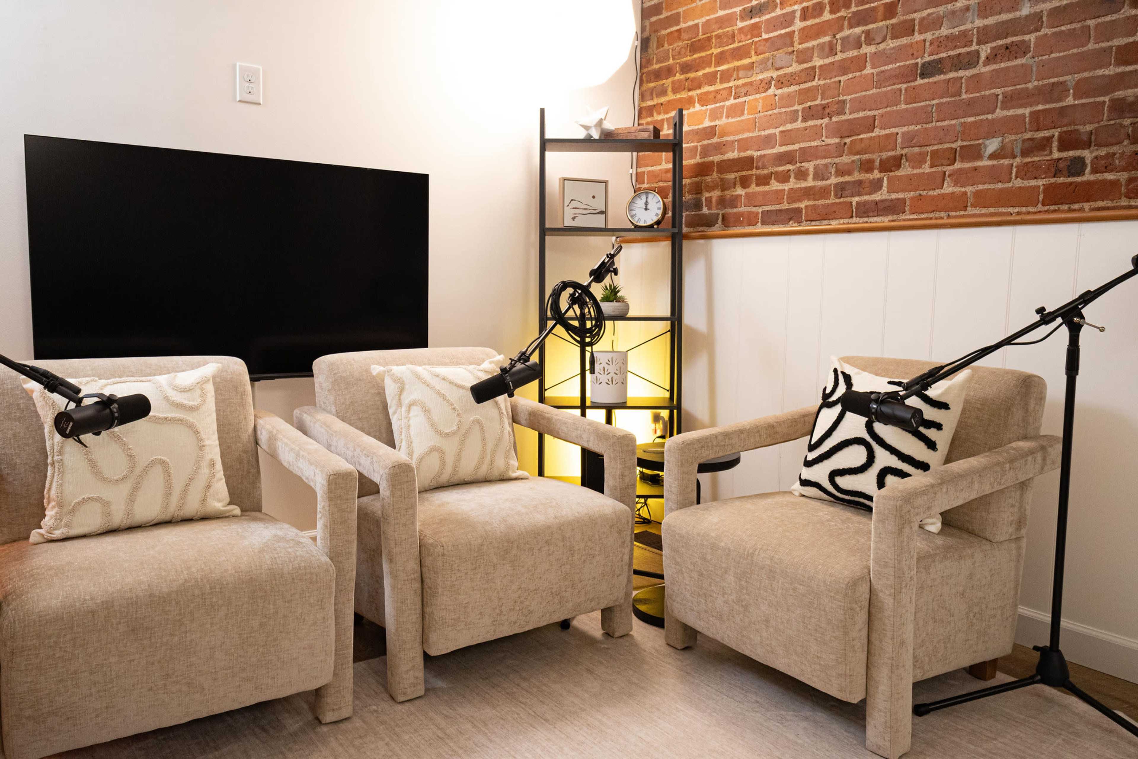 The image shows two beige armchairs with decorative pillows, positioned in front of a black screen and a shelf with various items, including a clock and a plant.