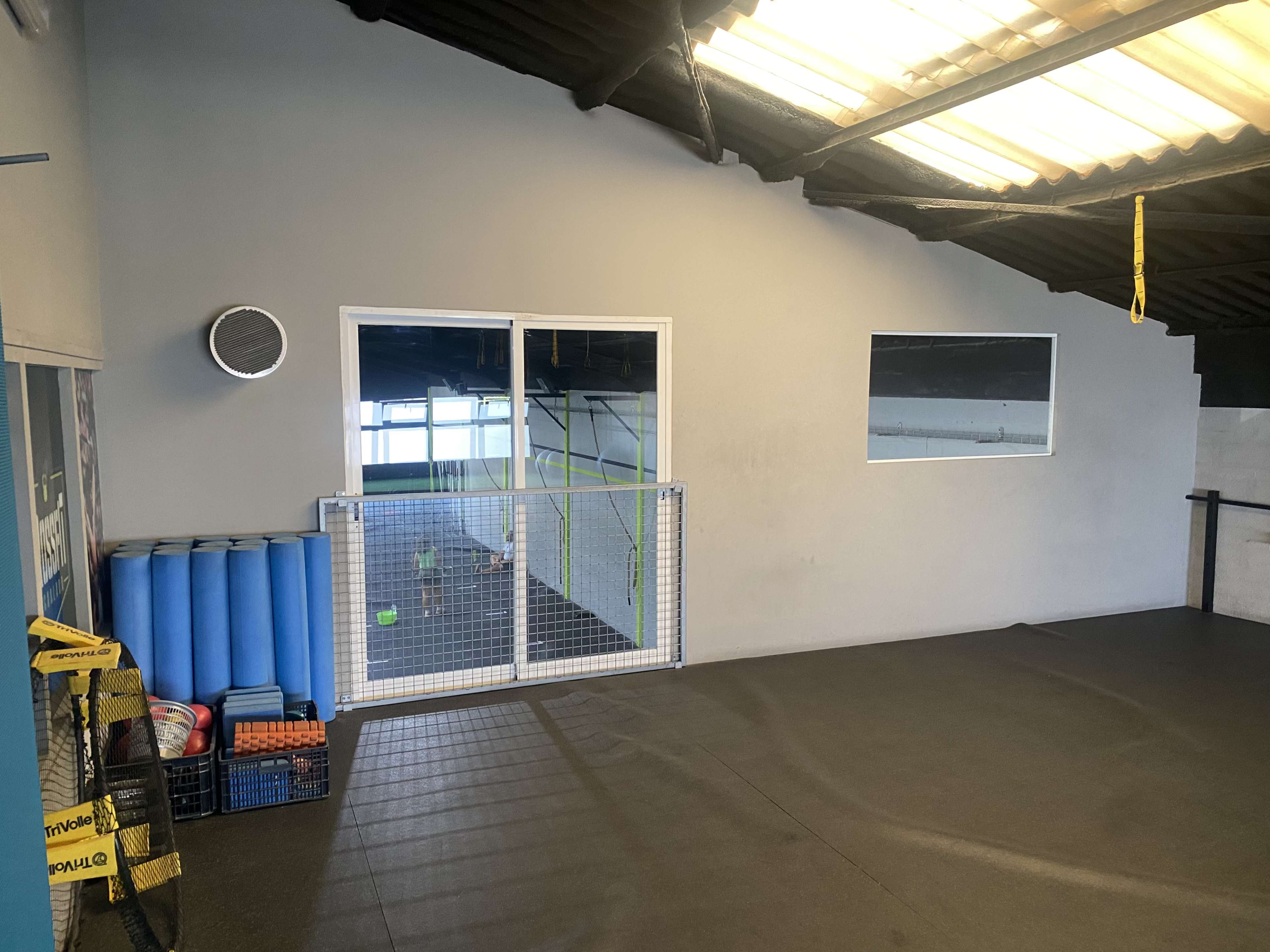 The interior of a gym features a mirrored wall, exercise mats stacked on one side, and a gated area with fitness equipment visible beyond a window.
