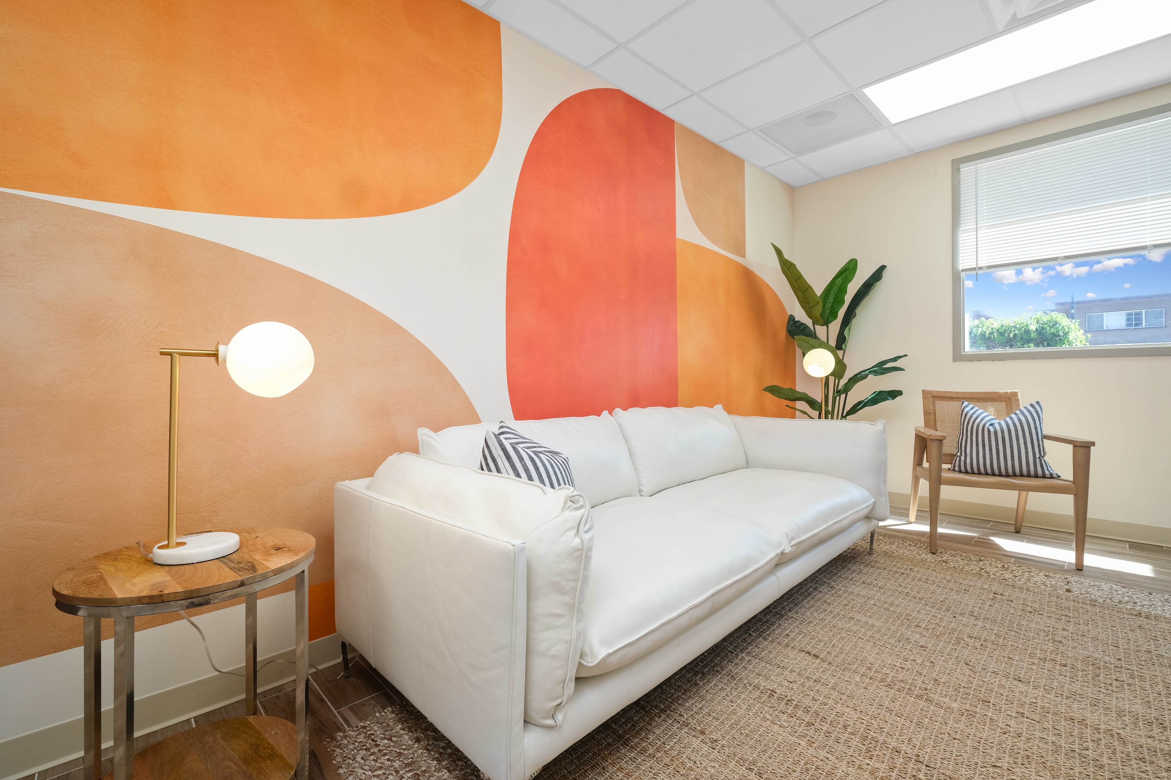 A light-colored couch in a room with a wall featuring abstract orange and peach shapes, accompanied by a plant and a small wooden table with a lamp.