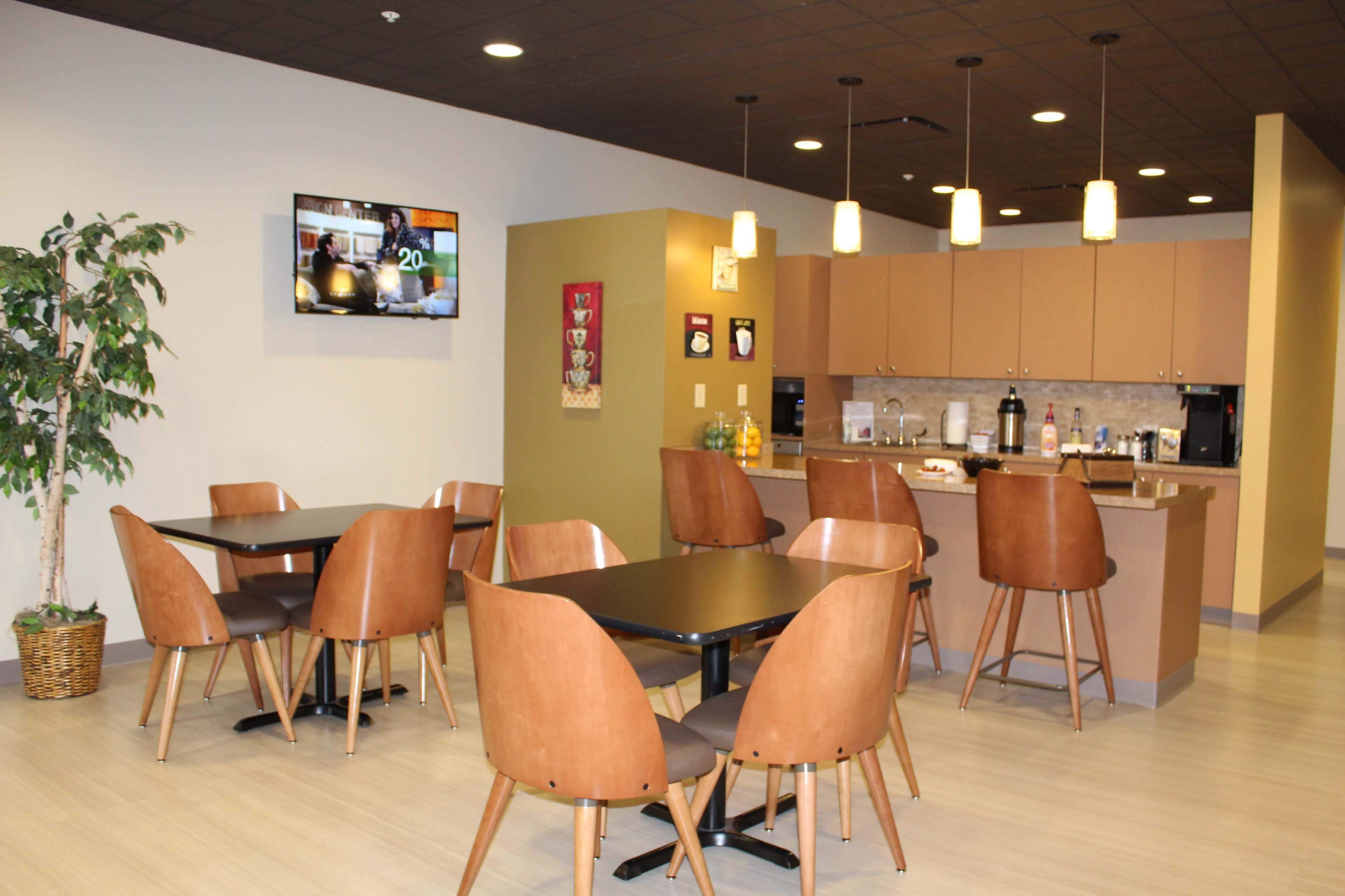 A modern dining area with brown chairs and tables, a kitchenette in the background, and a television mounted on the wall.