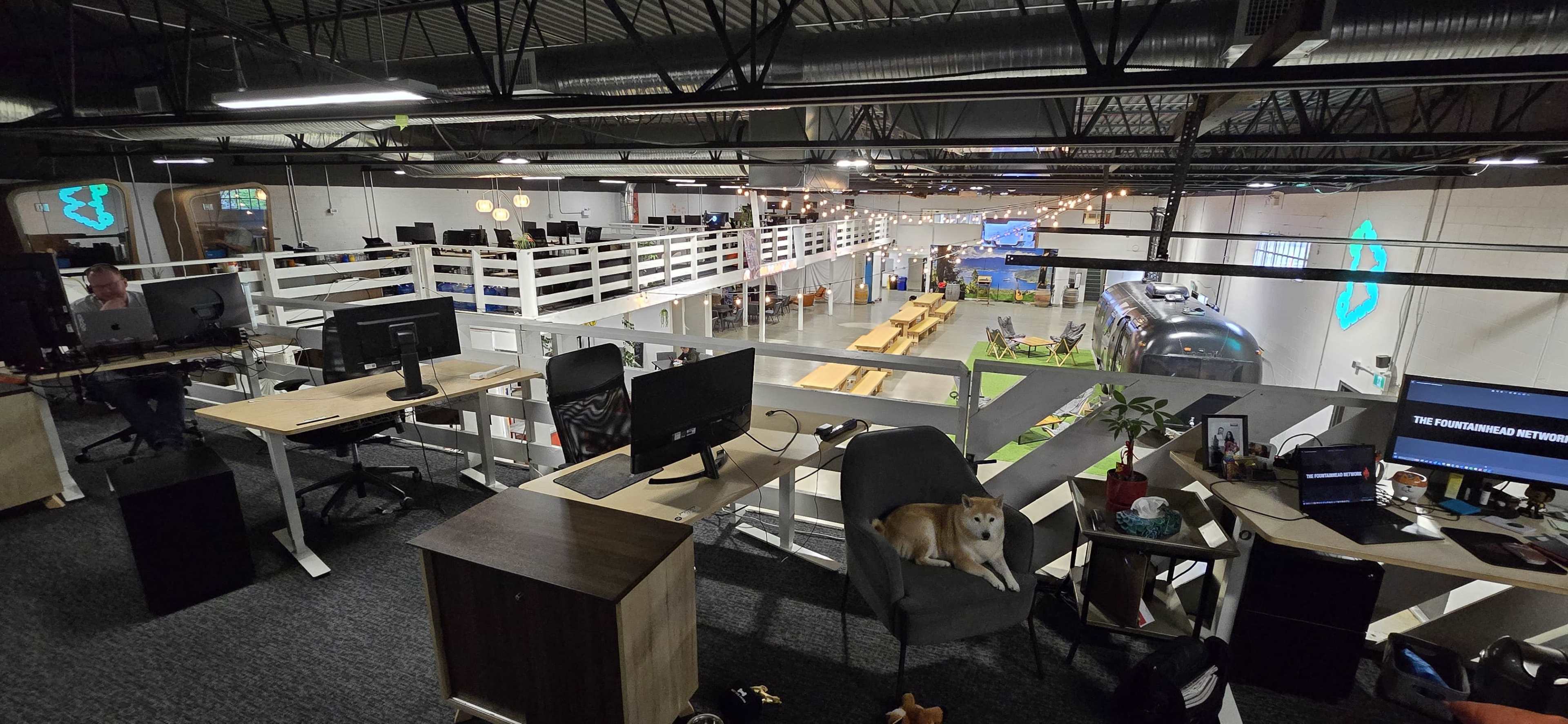 An open office space with multiple workstations, a suspended seating area, and a common dining area featuring a vintage Airstream trailer.