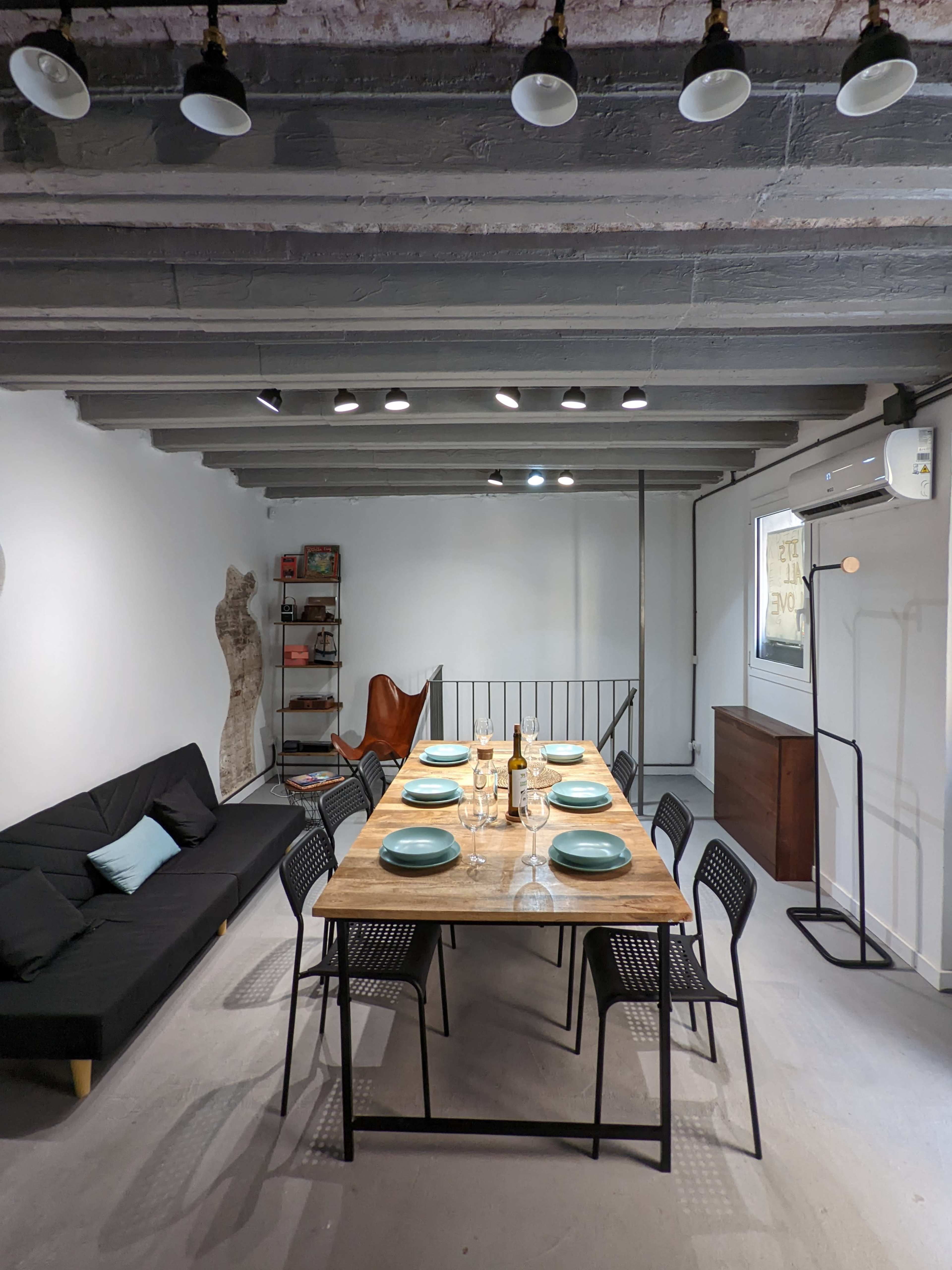 The image shows a modern dining area featuring a wooden table set for six, with black chairs, a black sofa, and artwork on the walls.