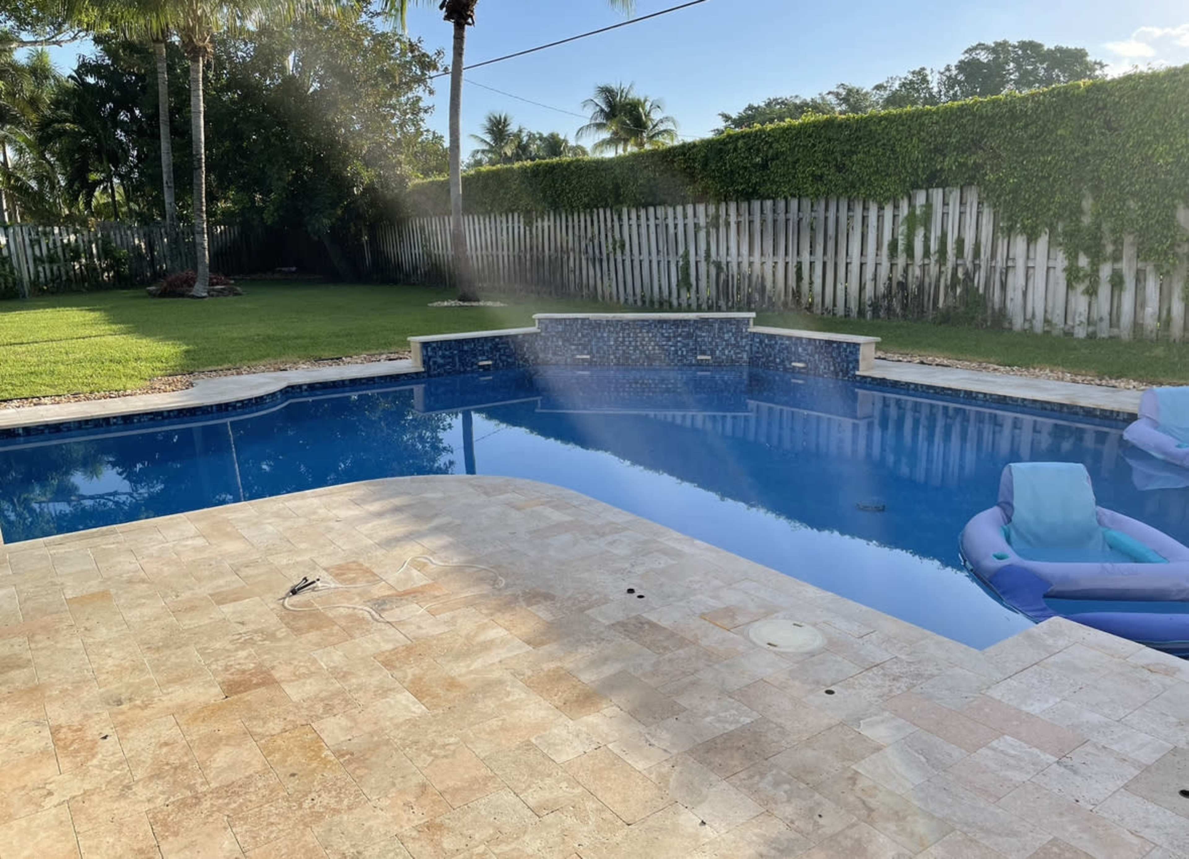 A pool with a stone deck features a blue tiled wall at one end, surrounded by a grassy area and palm trees.