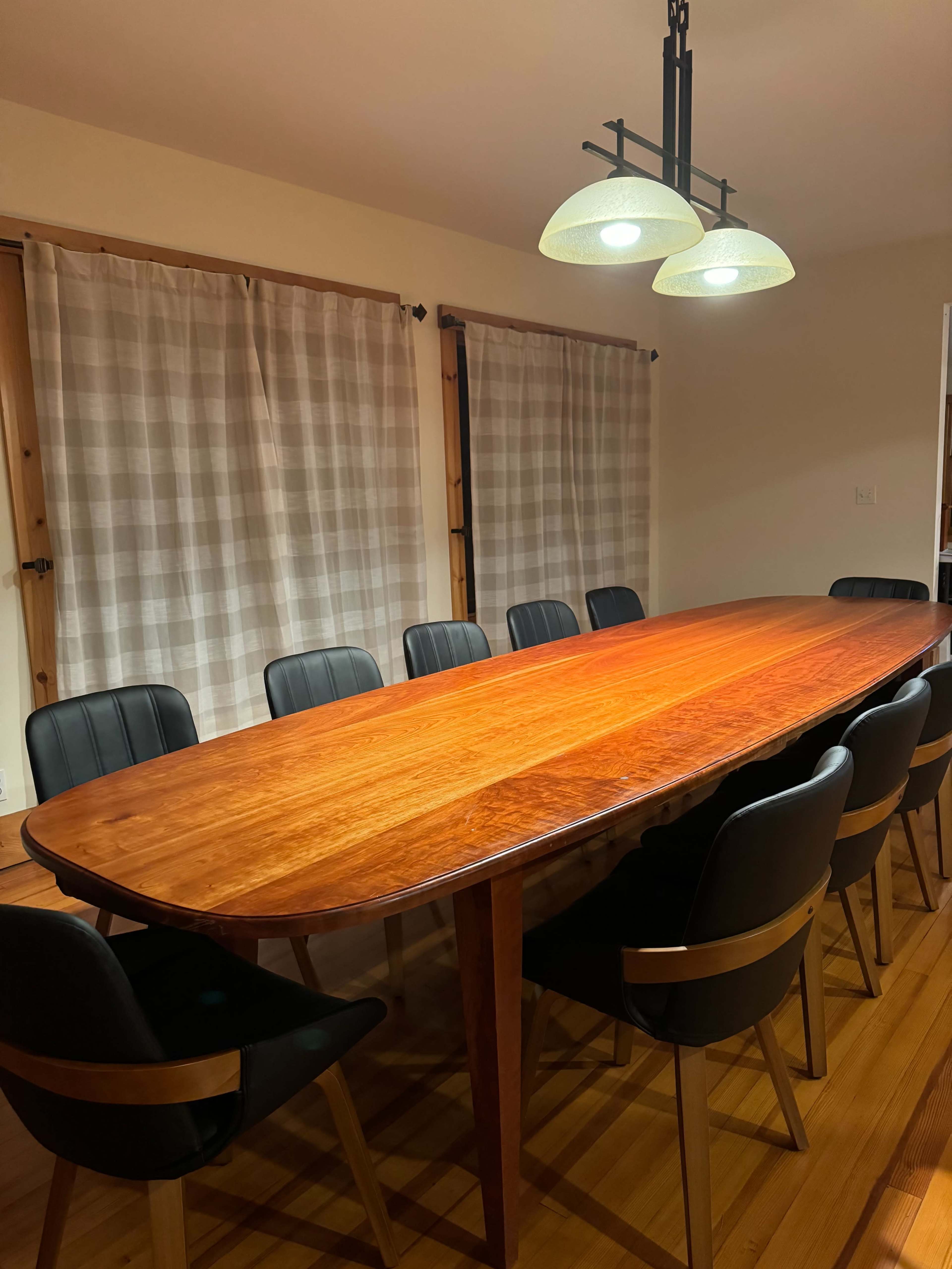 A large wooden dining table is surrounded by eight black chairs in a well-lit room with two curtain-covered windows.