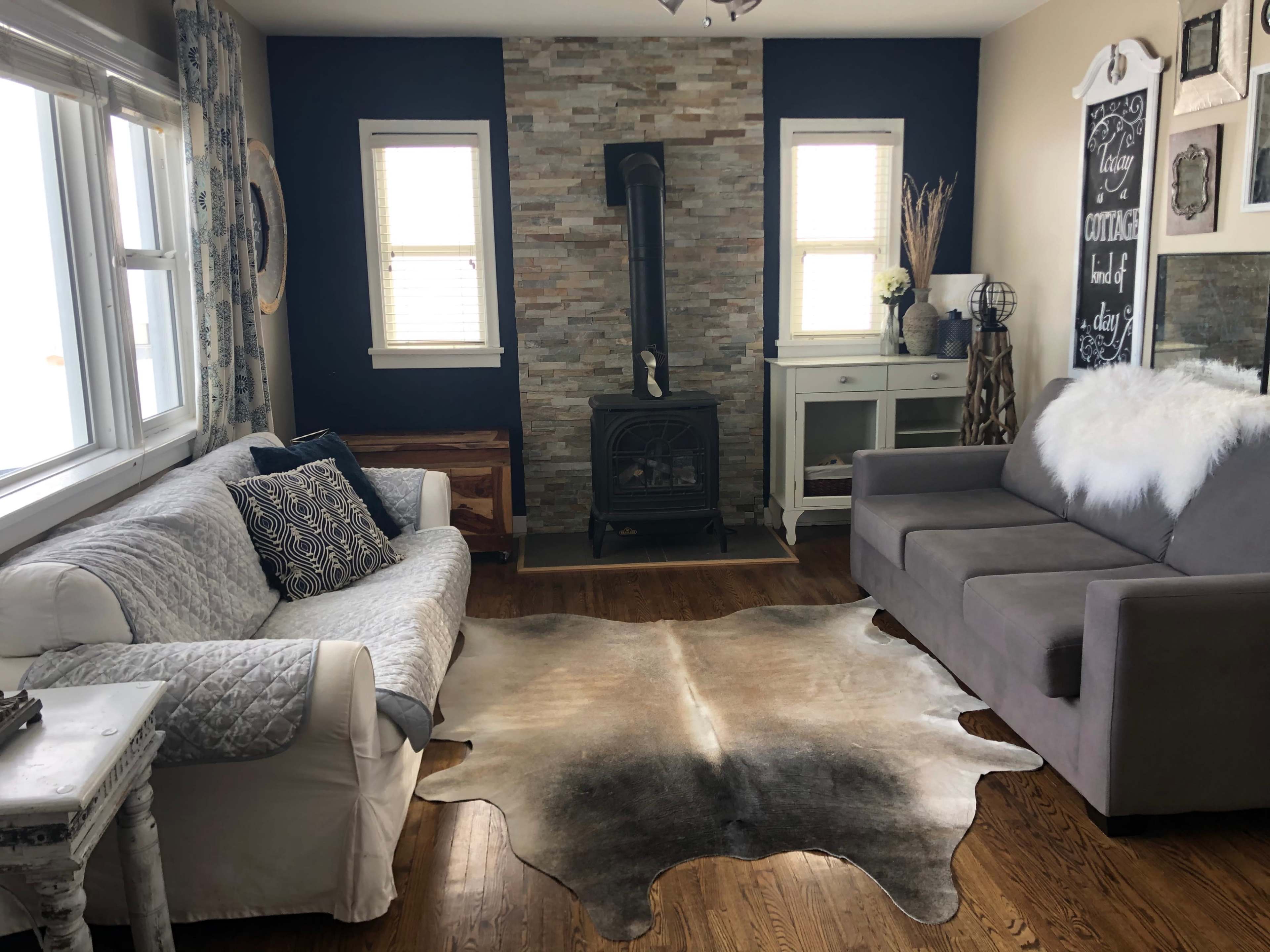 The image shows a living room with two sofas, a stone fireplace in the center, and a cowhide rug on a hardwood floor.
