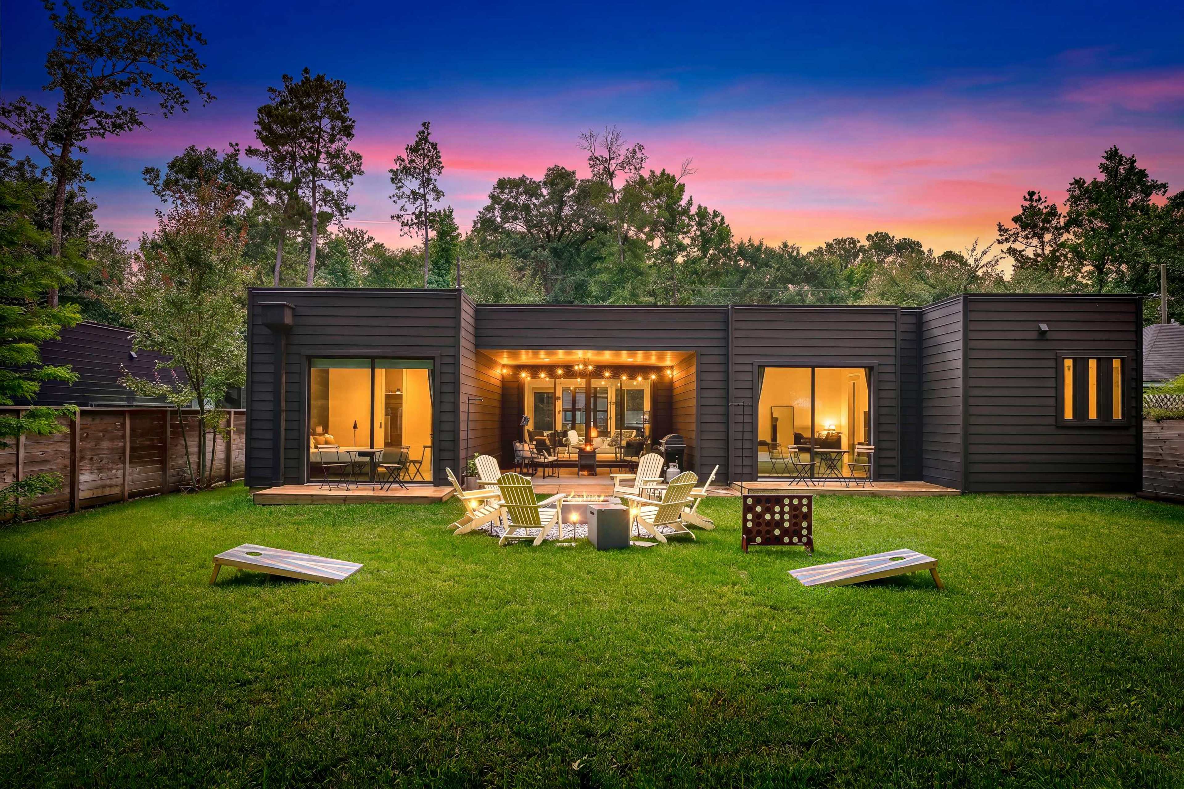 A modern black house with large windows sits in a green yard, surrounded by trees and outdoor seating under a colorful sunset.