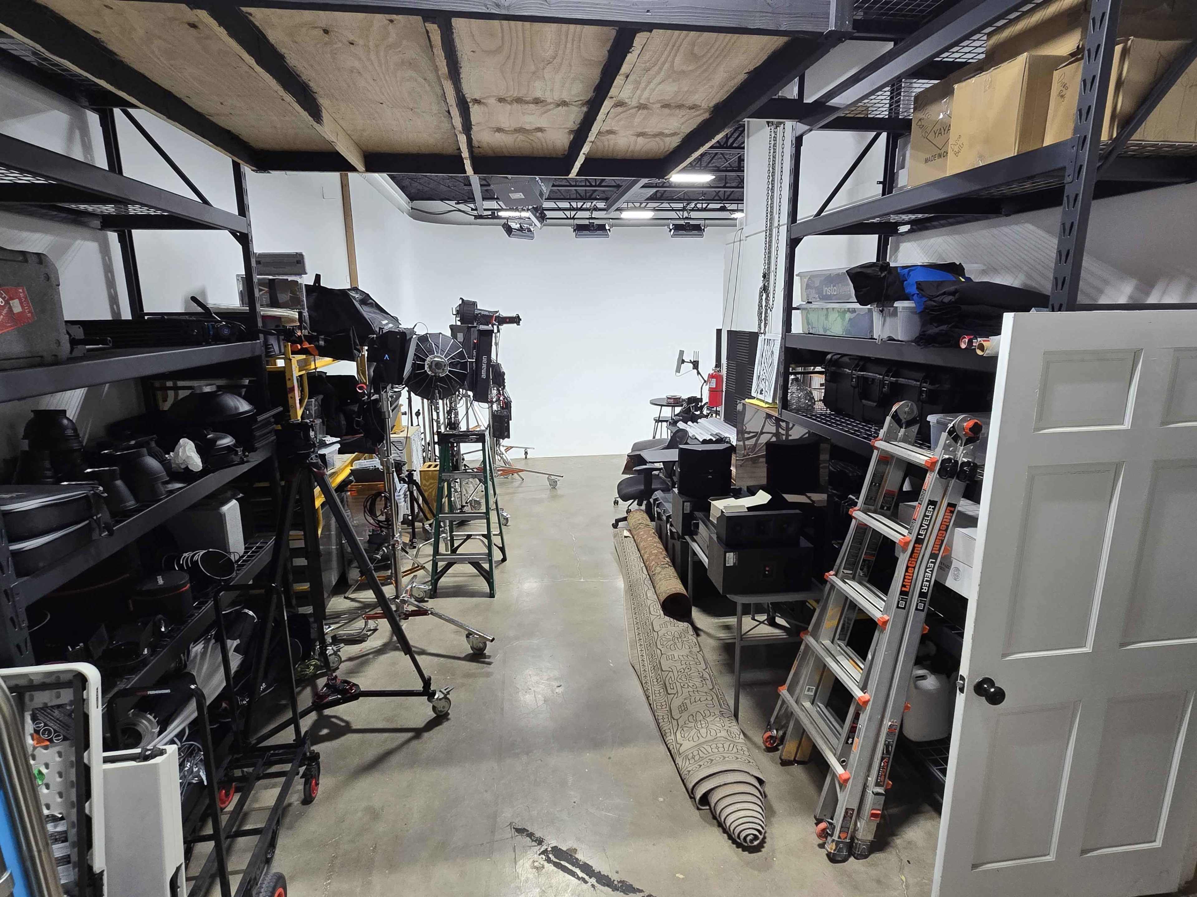 The image shows a cluttered photography studio space with shelves filled with equipment and props on either side, leading to a blank white wall at the end.