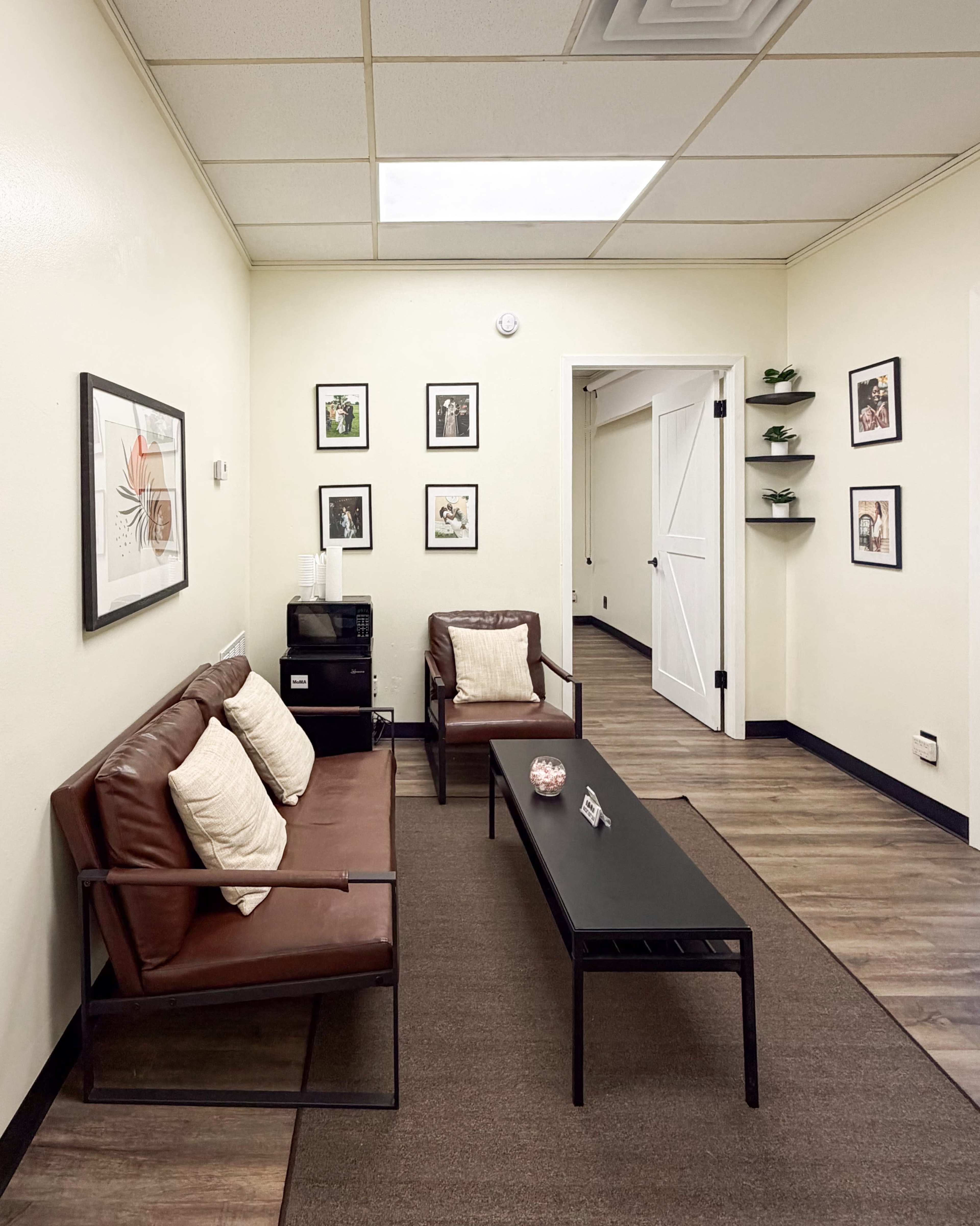 The image shows a small waiting area with two brown leather chairs and a coffee table, framed photographs on the walls, and a shelf with plants.