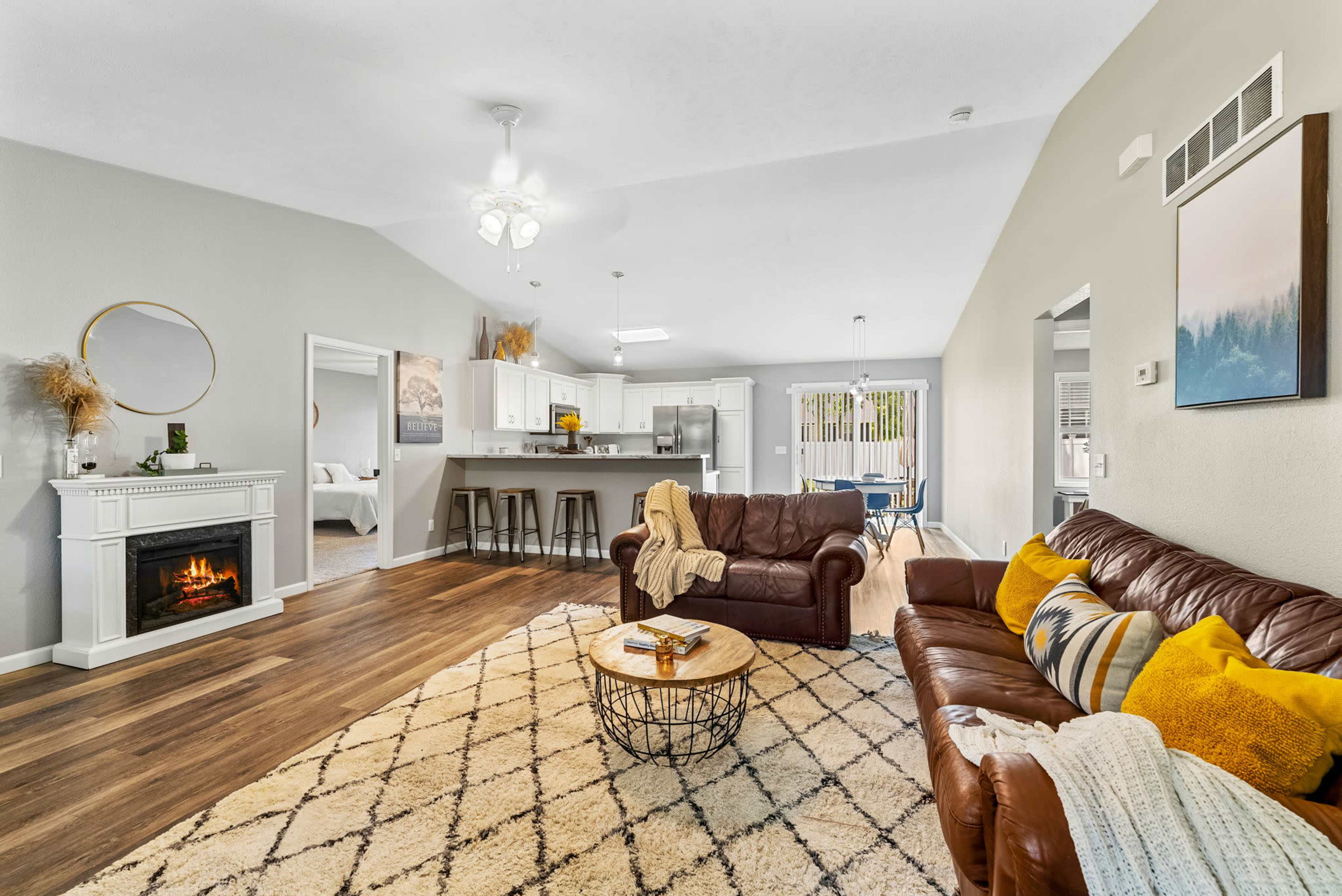 The living room features a brown leather sofa and an adjacent round coffee table, with a fireplace and an open layout leading to a modern kitchen.