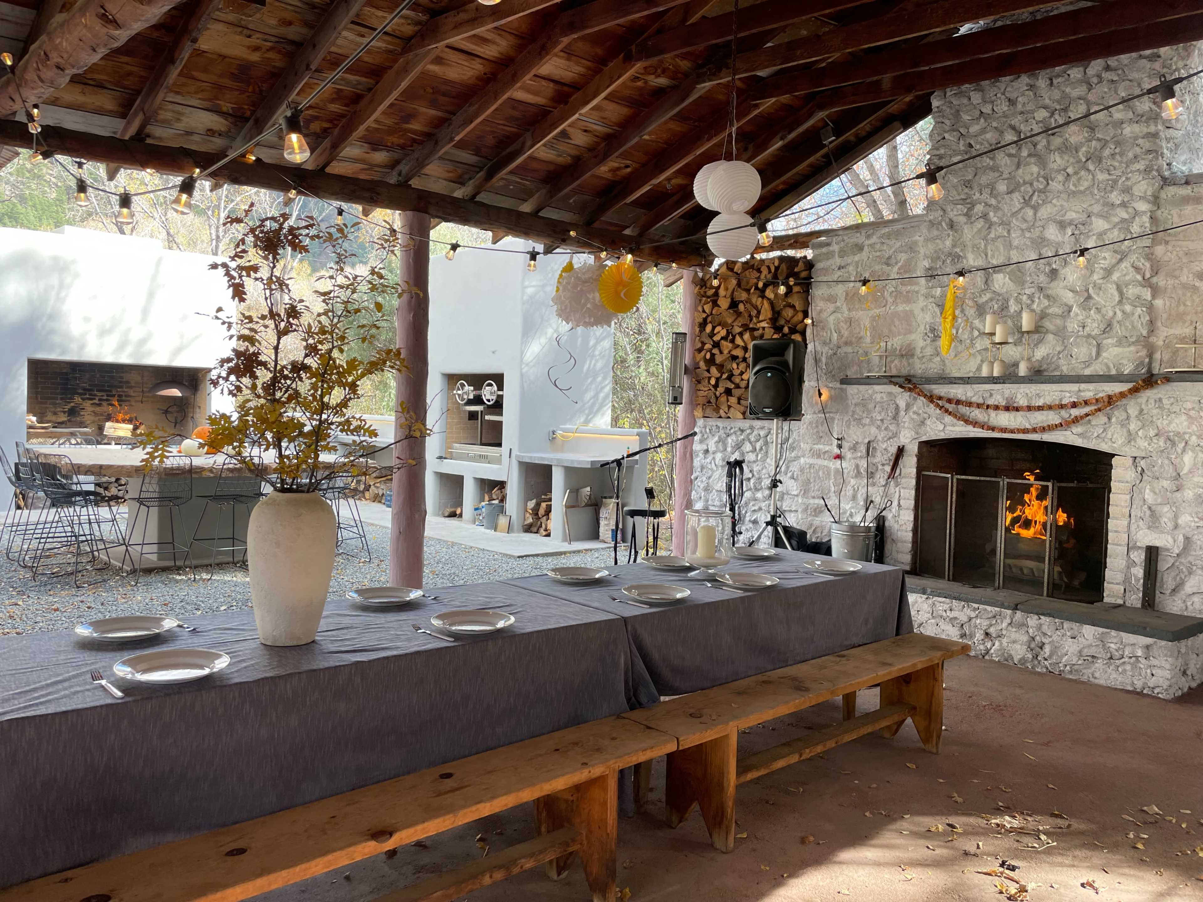 A rustic outdoor dining area under a wooden roof, featuring a long table with plates, a stone fireplace, and a gathering space decorated with lights and decorations.