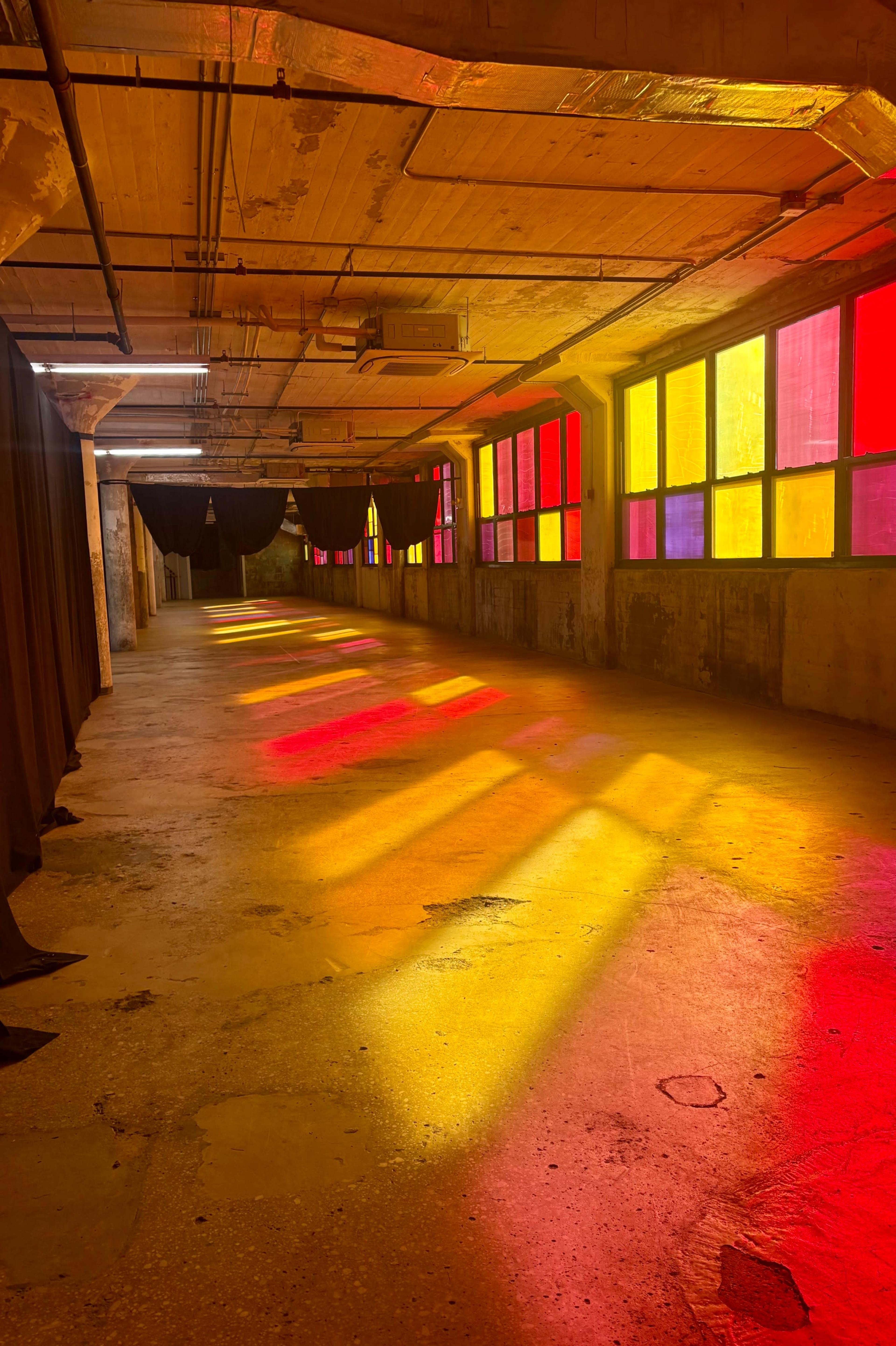 A long, empty hallway features colorful light patterns projected through stained glass windows onto the floor.