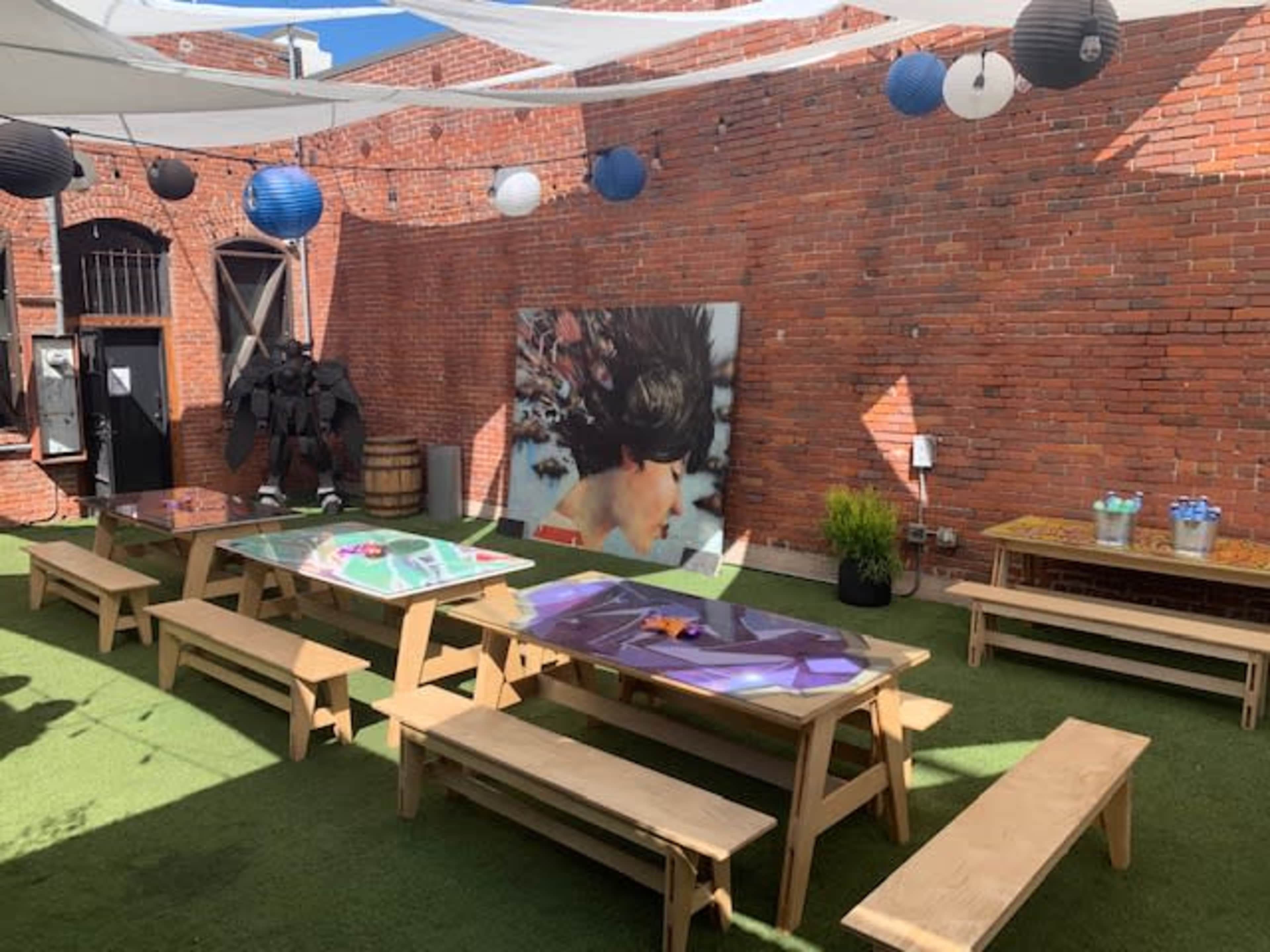 The scene features an outdoor space with wooden benches and tables under a canopy, surrounded by brick walls and decorated with colorful lanterns and a large mural.