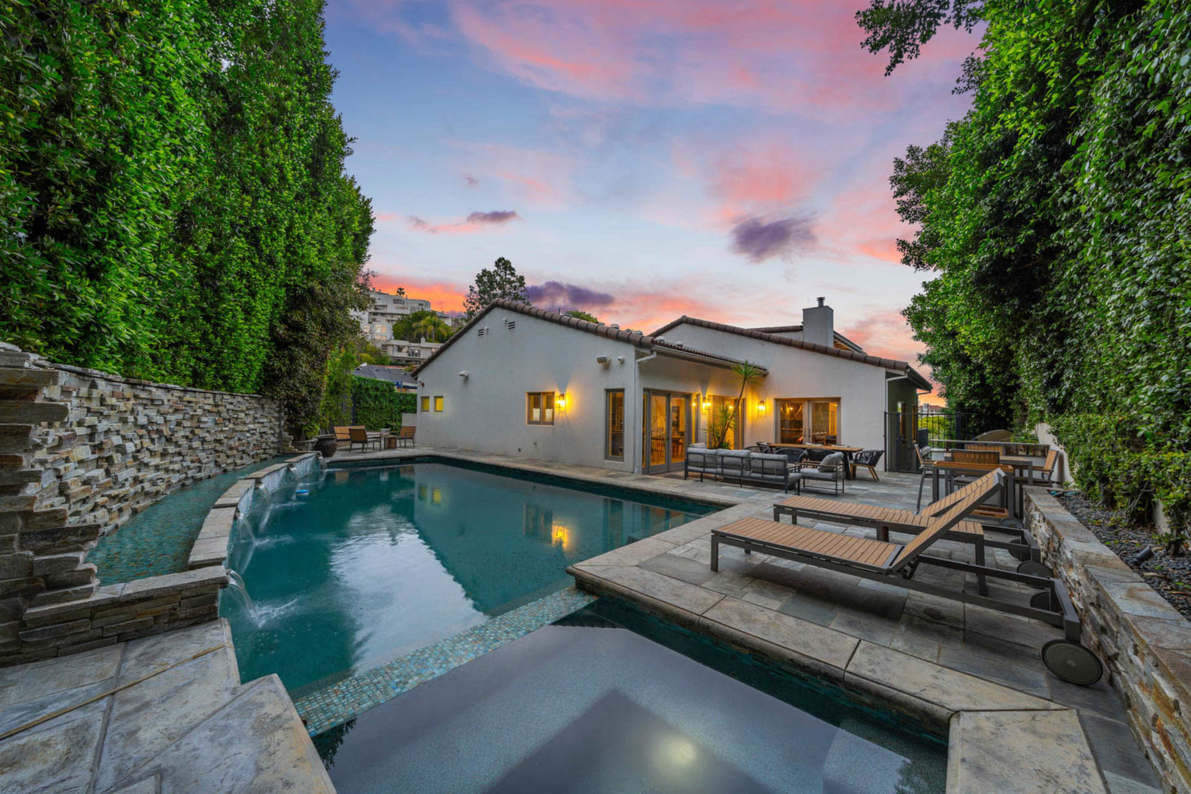 A modern home with a swimming pool is surrounded by lush greenery and features a scenic sunset in the background.