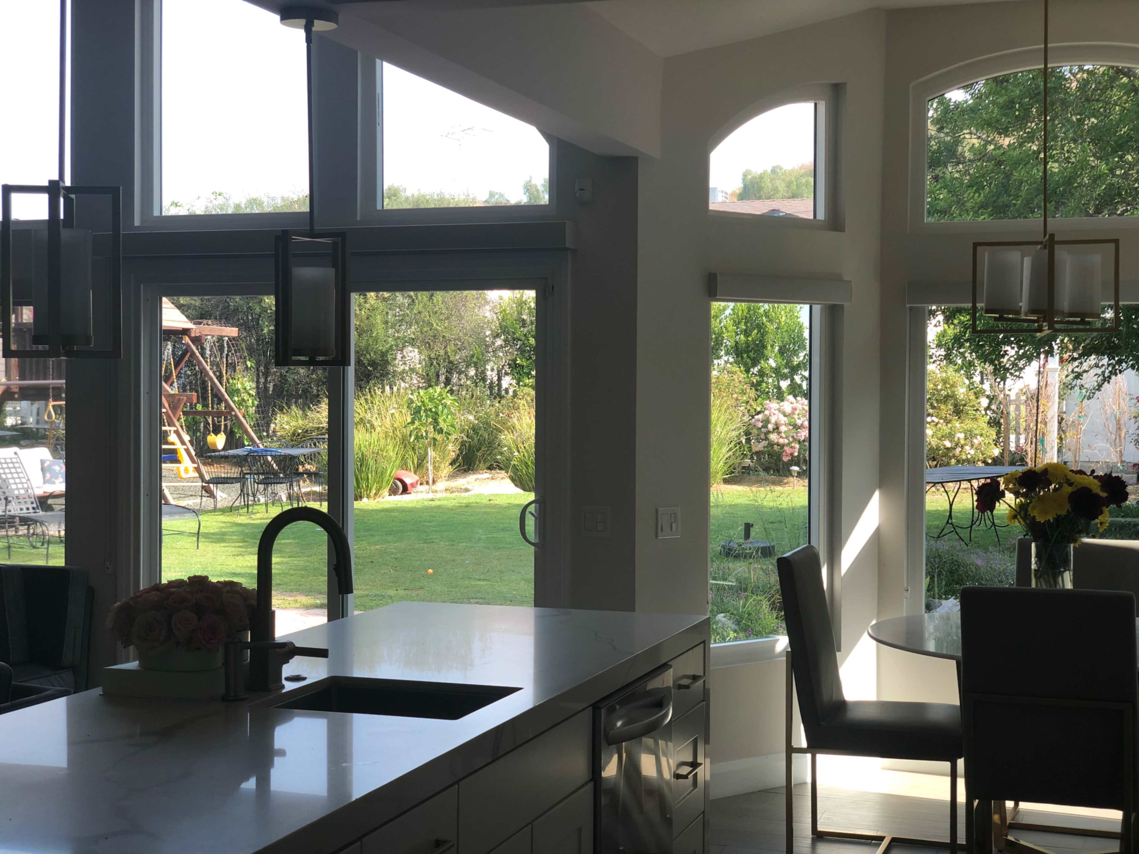 A modern kitchen with a large island overlooks a backyard featuring greenery and a swing set through expansive windows.