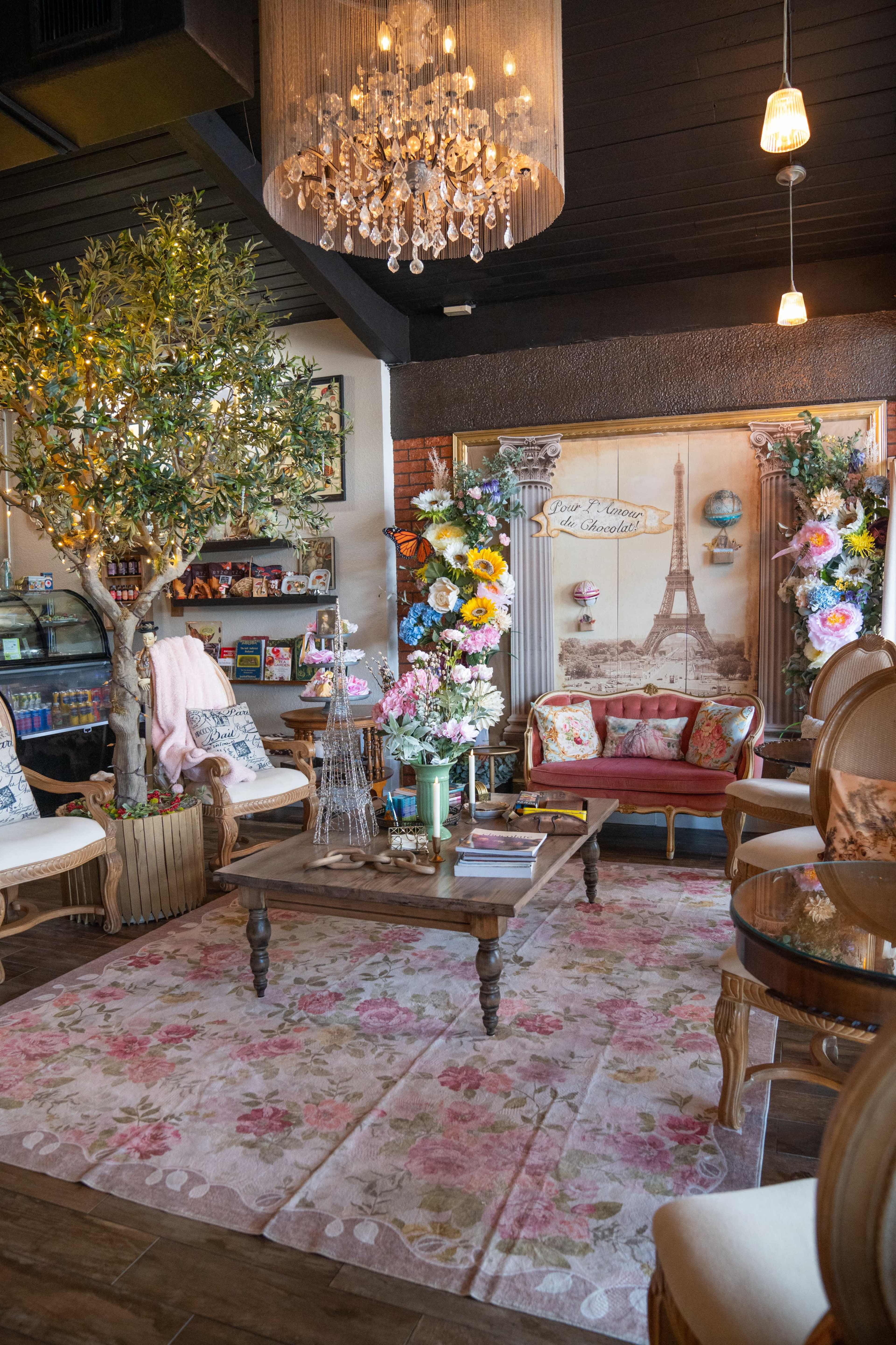 The image shows a cozy café interior with floral decorations, a chandelier, and vintage furniture, including a table surrounded by chairs.
