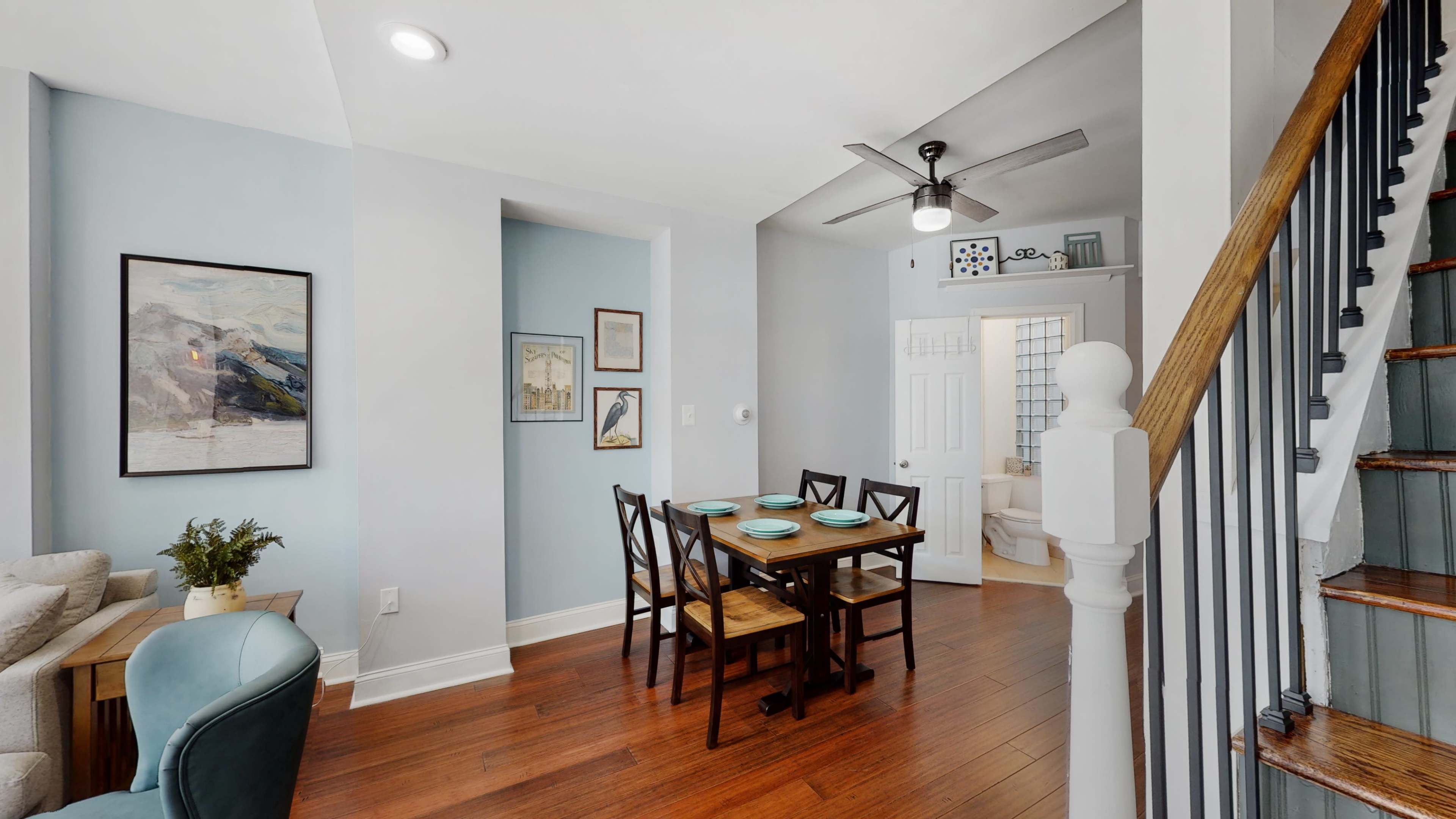 A well-lit dining area featuring a table with four chairs, surrounded by light blue walls and hardwood flooring, with a staircase visible in the background.