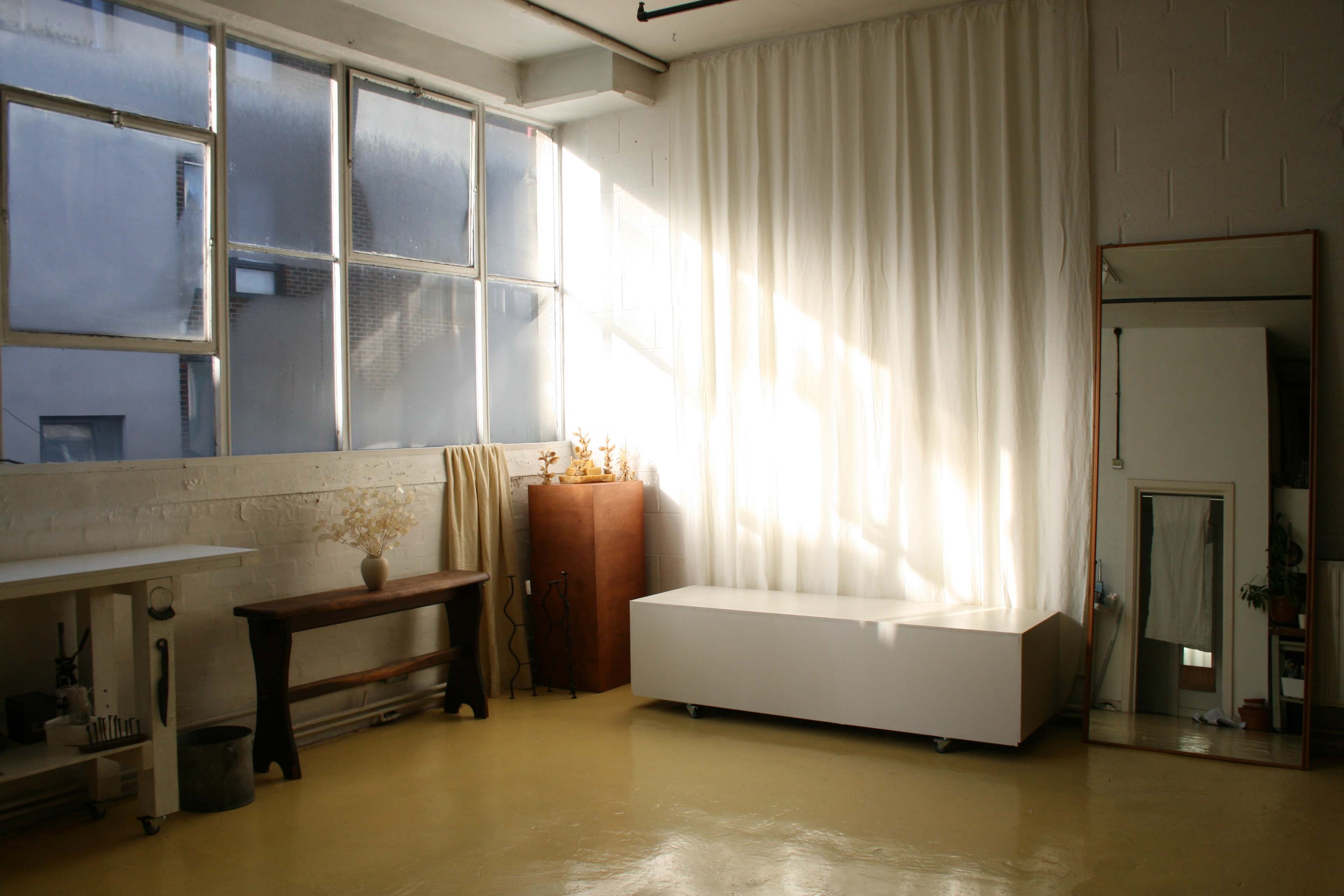 A bright, minimalistic studio features large windows, a white curtain, a wooden table, a modern white cabinet, and a tall mirror against the wall.