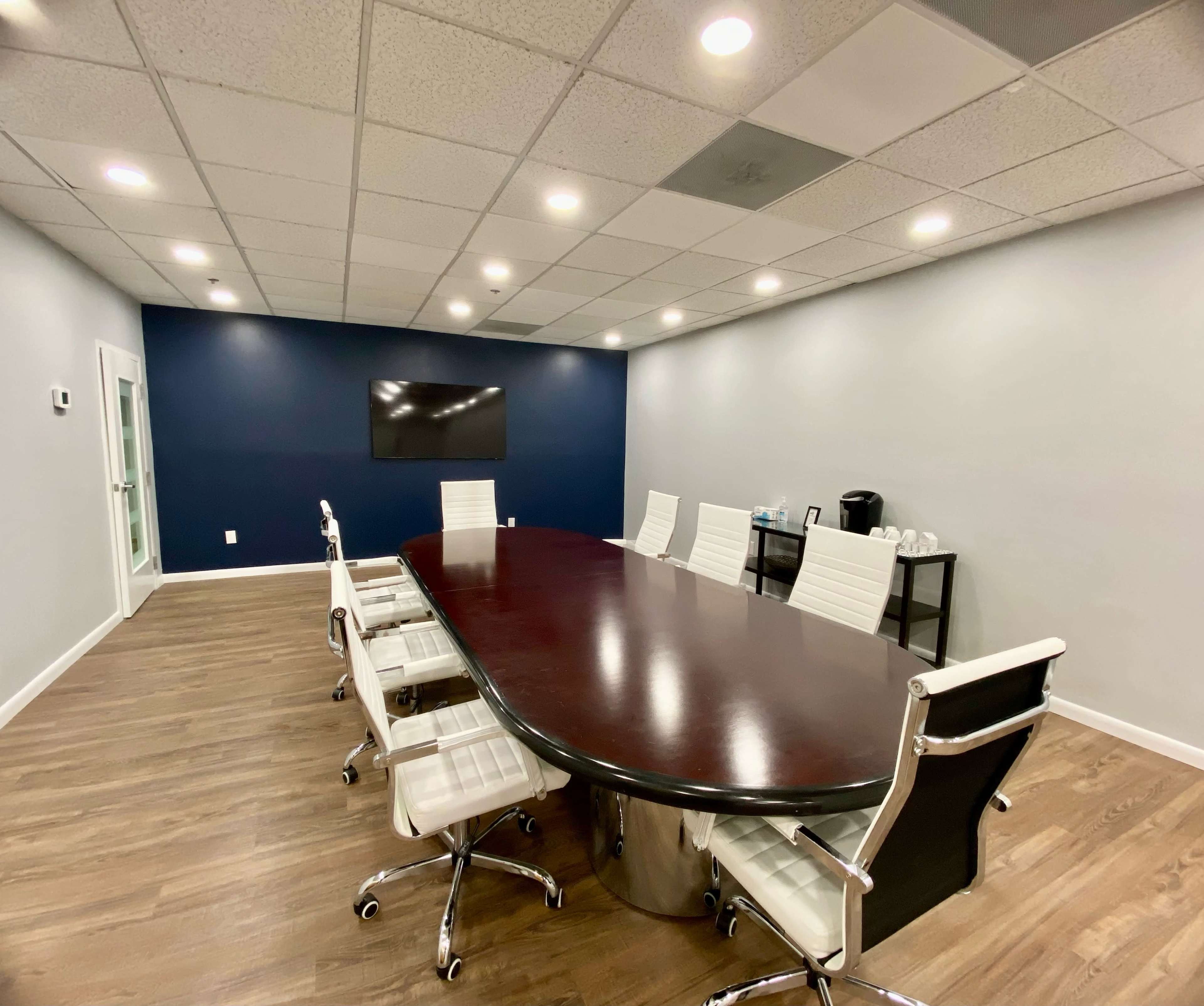 A large conference room features a long wooden table surrounded by white chairs, with a television mounted on the wall and a coffee station in the corner.