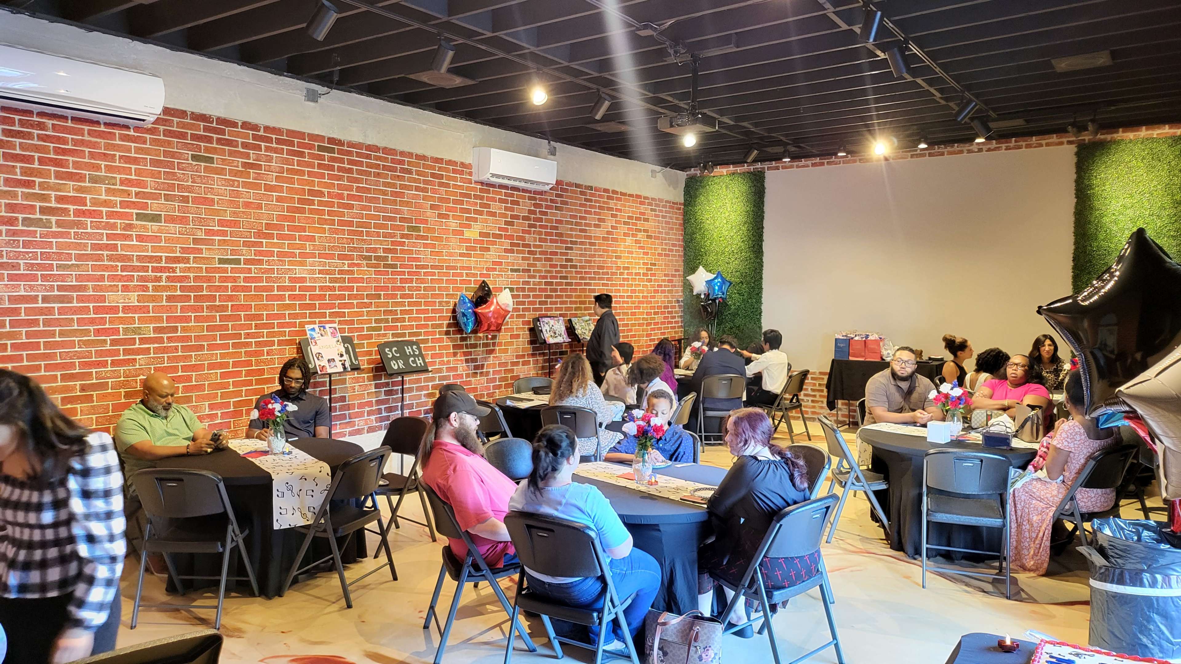 The image shows a crowded event space with tables arranged for guests, featuring a brick wall and decorative greenery.